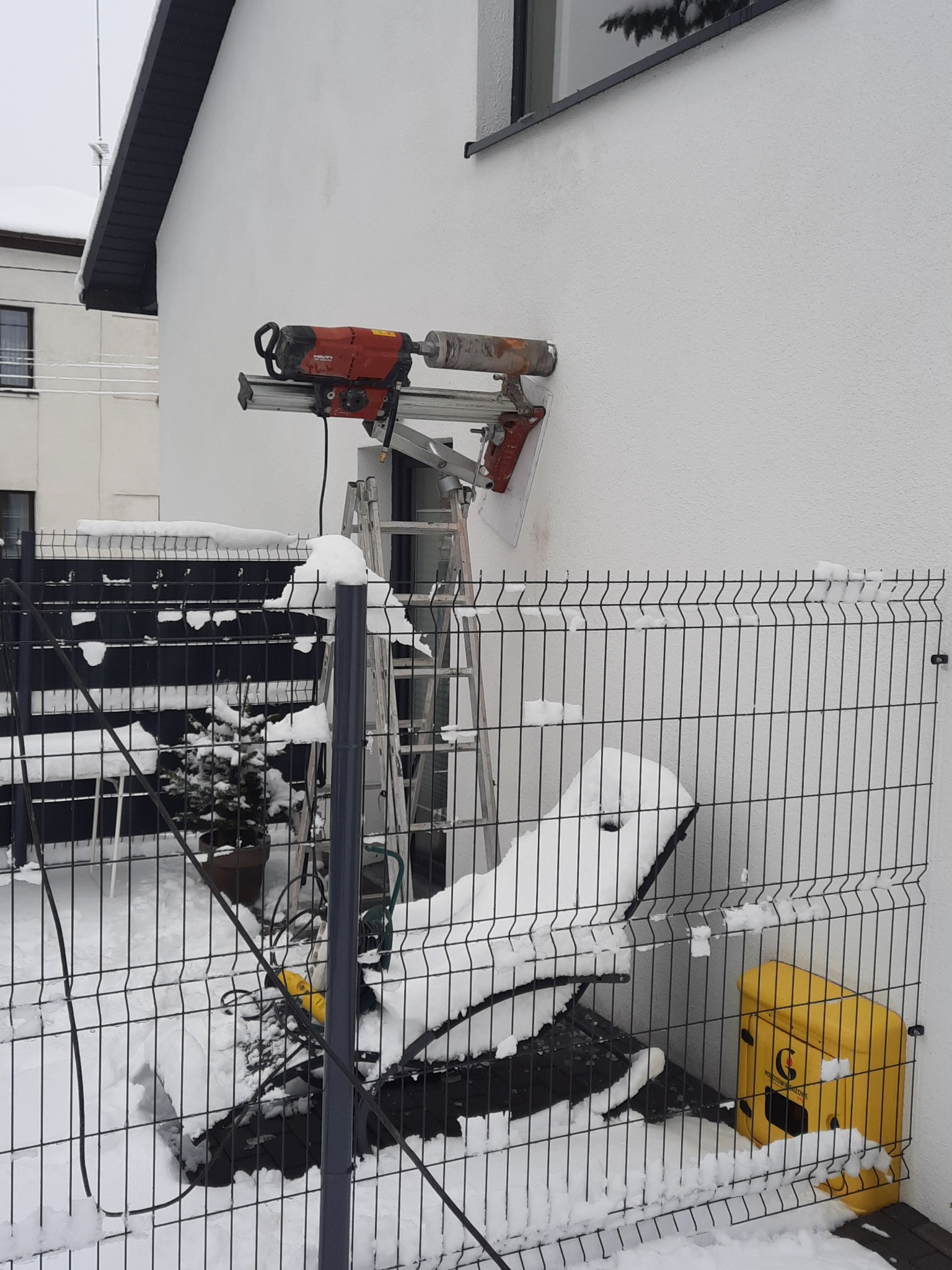 Czerwona wiertnica diamentowa Hilti zamocowana na ścianie budynku z białym tynkiem, wsparta na aluminiowej konstrukcji i drabinie w zimowej scenerii, widok przez metalowe ogrodzenie pokryte śniegiem.