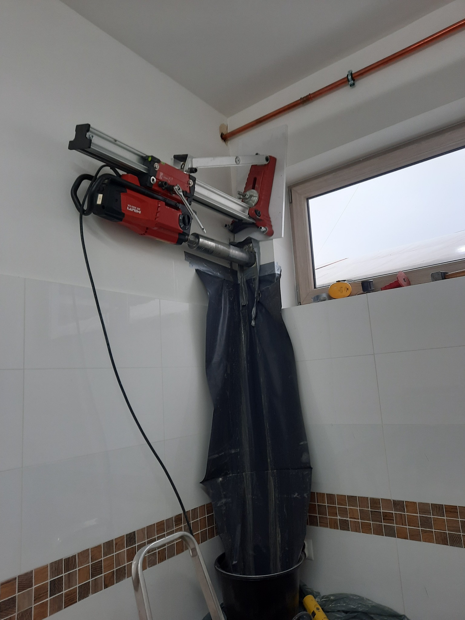 Czerwona wiertnica diamentowa Hilti zamocowana do ściany z biało-brązowymi płytkami, wiercąca otwór; czarny worek zbiera urobek, widoczna rura miedziana pod sufitem i drabina w tle.