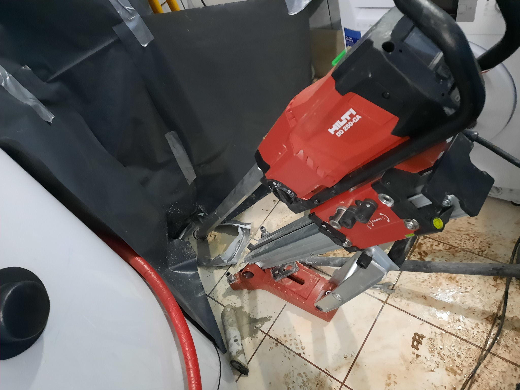 Czerwona wiertnica Hilti DD 250-CA w trakcie pracy, wiercenie rdzeniowe w podłodze z płytek, widoczny wąż doprowadzający wodę, czarna folia ochronna i resztki po wierceniu.
