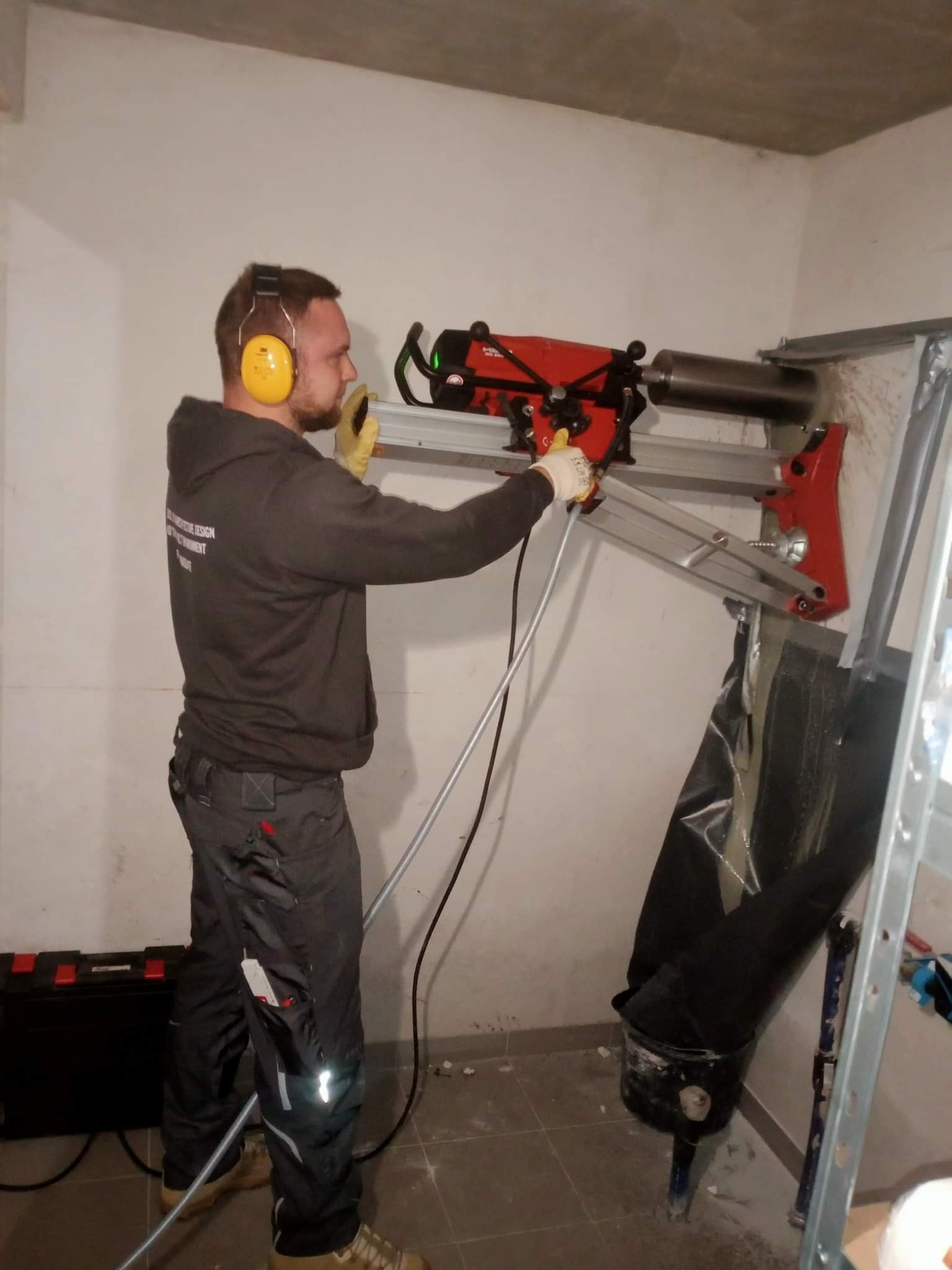 Pracownik w słuchawkach ochronnych wierci otwór w ścianie za pomocą wiertnicy diamentowej z odsysaniem urobku, widoczna skrzynka narzędziowa i fragment ściany z ubytkiem.