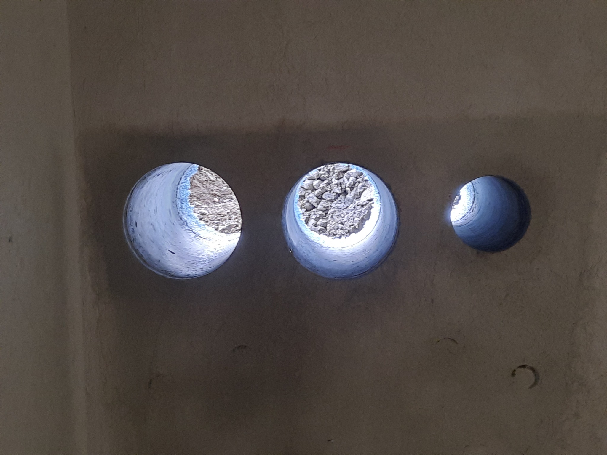 Trzy otwory wywiercone w szarej ścianie, o różnych średnicach, ukazujące gruz i ziemię wewnątrz, z widocznymi śladami wiercenia.