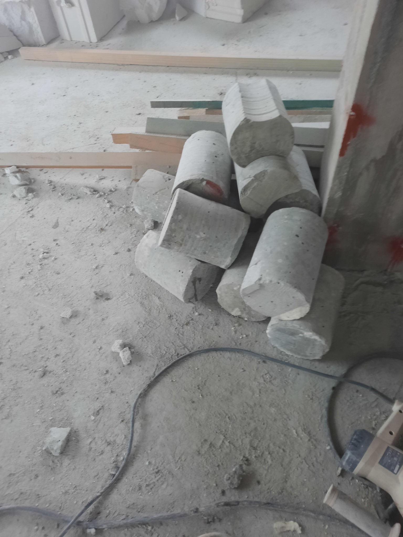 Rdzenie betonu ułożone na szarym, zakurzonym podłożu, w pobliżu wiertarki rdzeniowej i drewnianych elementów konstrukcyjnych.