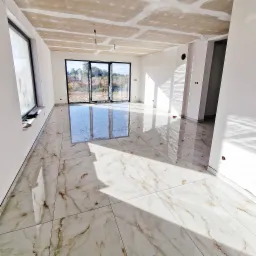 Salon w stanie deweloperskim z ułożoną, błyszczącą podłogą z płytek imitujących marmur, surowy sufit z widocznymi łączeniami płyt gipsowo-kartonowych, duże okna balkonowe z widokiem na zewnątrz.