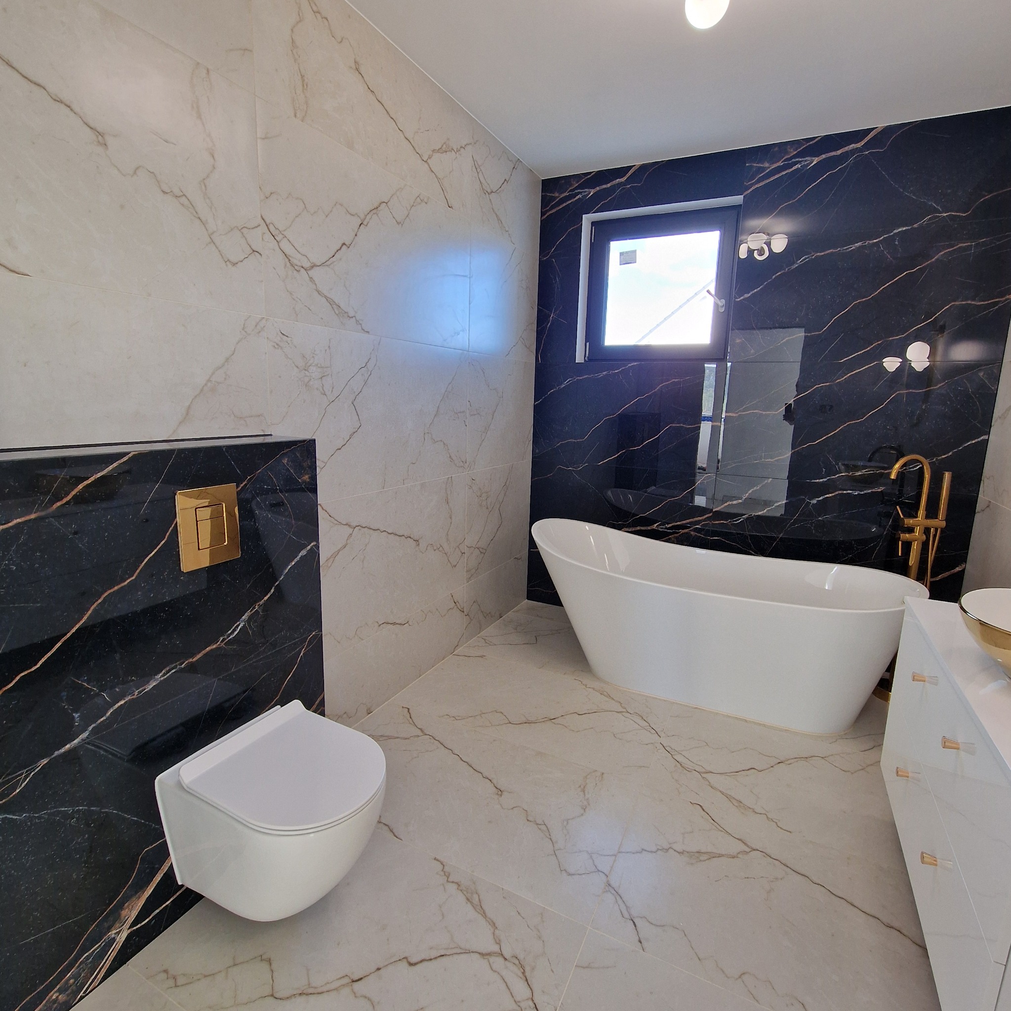 Elegancka łazienka z białymi sanitariatami i wanną wolnostojącą, wykończona płytkami imitującymi marmur w odcieniach bieli i czerni z złotymi akcentami.