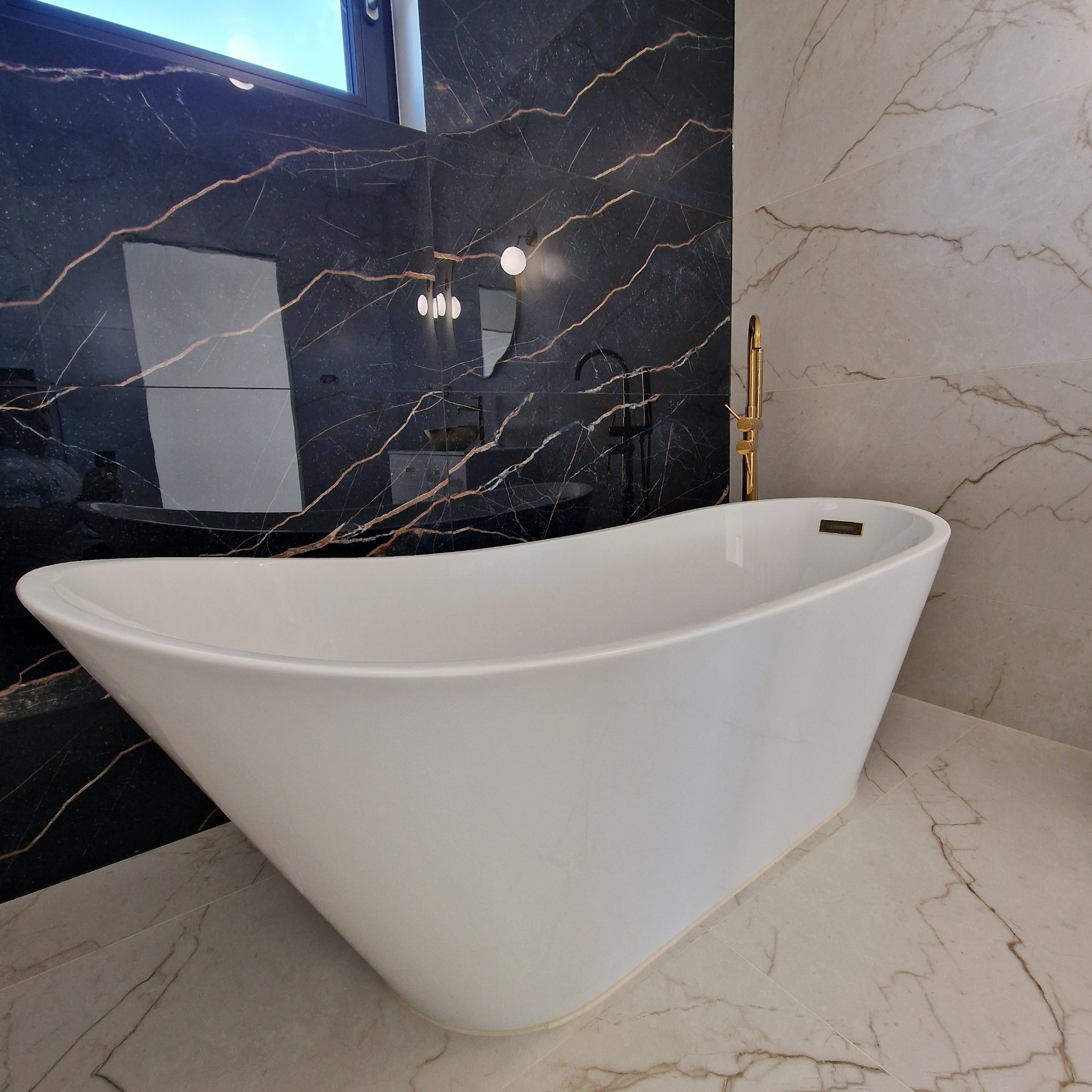 Wolnostojąca, biała wanna na tle ściany z czarnego marmuru z żyłkami i jasnego marmuru, złota bateria podłogowa