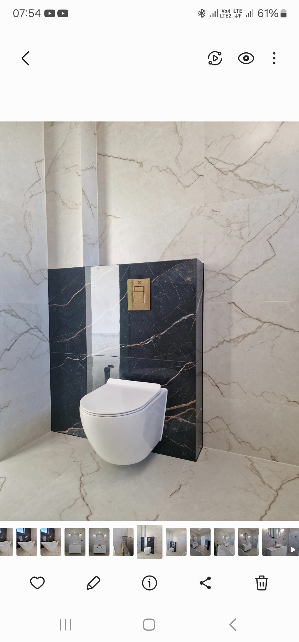 Podwieszana toaleta w nowoczesnej łazience z marmurowymi płytkami na ścianach i podłodze, czarny panel z złotym przyciskiem spłuczki.