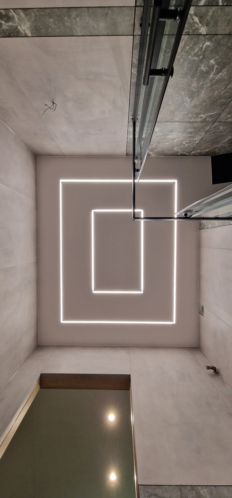 Nowoczesna łazienka z szarymi płytkami, minimalistycznym oświetleniem LED w kształcie kwadratów na suficie i kabiną prysznicową z czarnymi profilami.