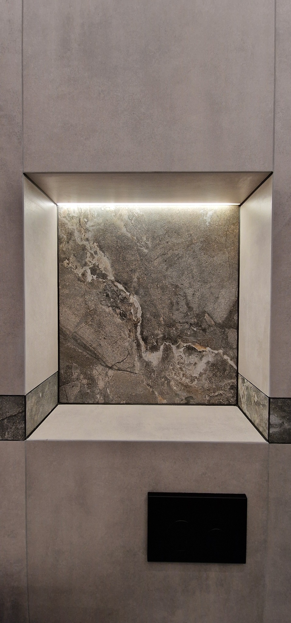 Płytki ścienne w łazience z wnęką podświetloną LED, wykończoną płytką imitującą marmur, z czarnym przyciskiem spłuczki.