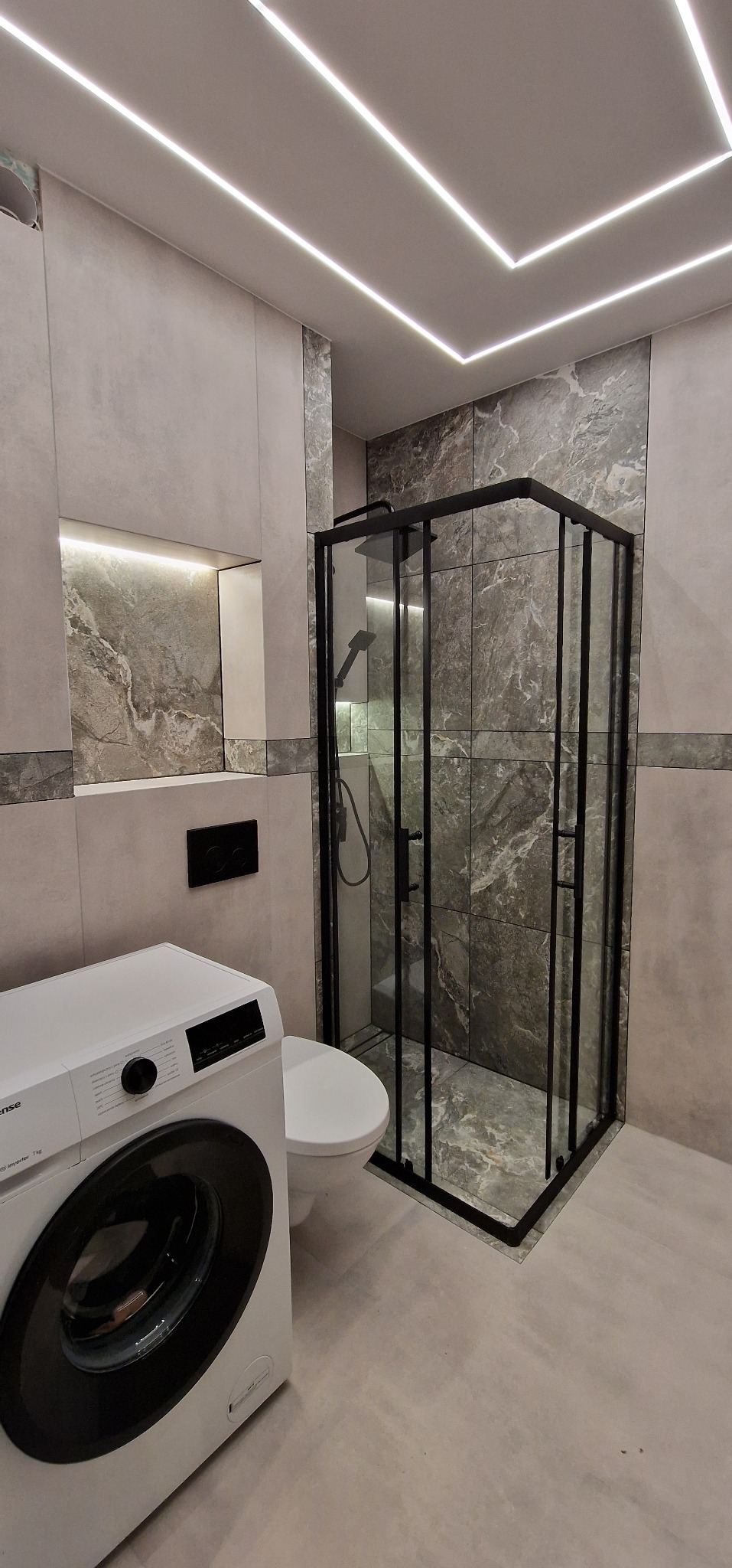 Nowoczesna łazienka z kabiną prysznicową w czarnej ramie, umywalką, toaletą i pralką Hisense, wykończona płytkami imitującymi marmur, z podświetlaną wnęką i liniowym oświetleniem sufitowym.