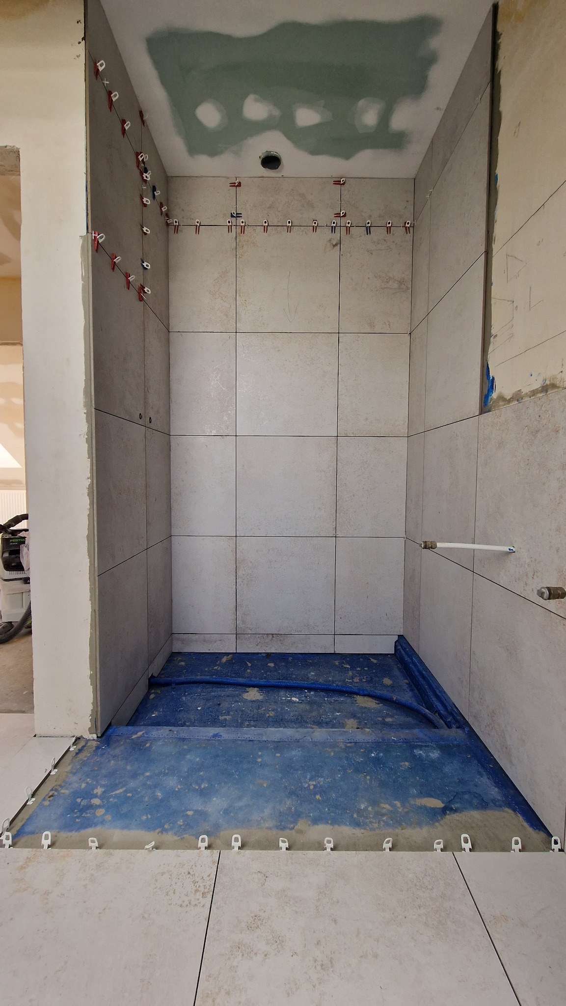 Montaż jasnych płytek w kabinie prysznicowej, widoczne systemy poziomowania płytek, niebieska folia wodoszczelna na podłodze, surowy stan wykończenia sufitu.