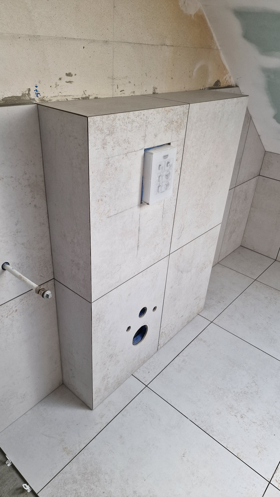 Ściana w łazience wyłożona jasnymi, kwadratowymi płytkami imitującymi kamień, z widocznymi otworami na instalacje i zamontowanym panelem sterowania, w trakcie prac wykończeniowych.