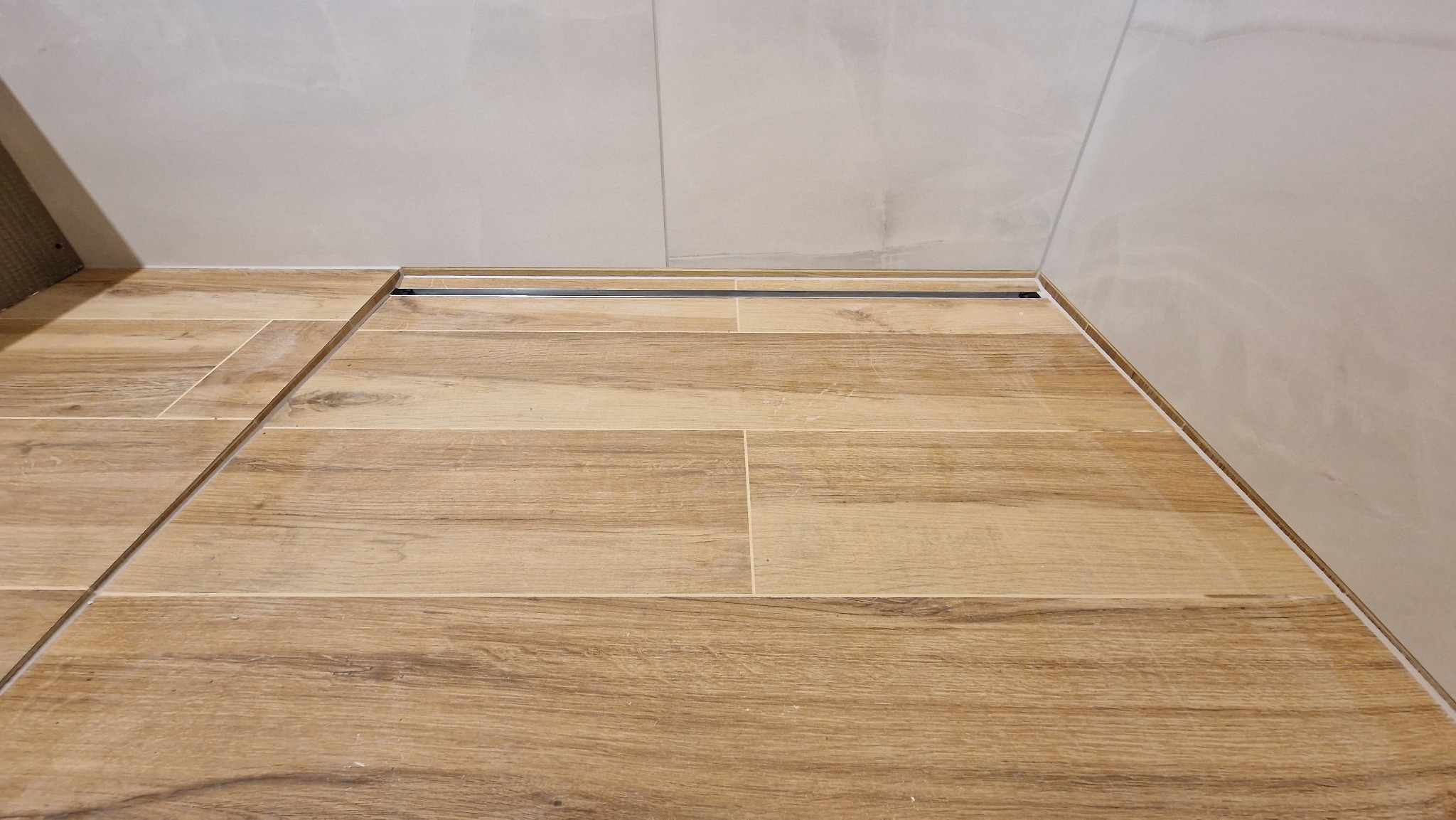 Ułożona podłoga z płytek imitujących drewno w łazience z odpływem liniowym przy ścianie wyłożonej jasnymi płytkami.