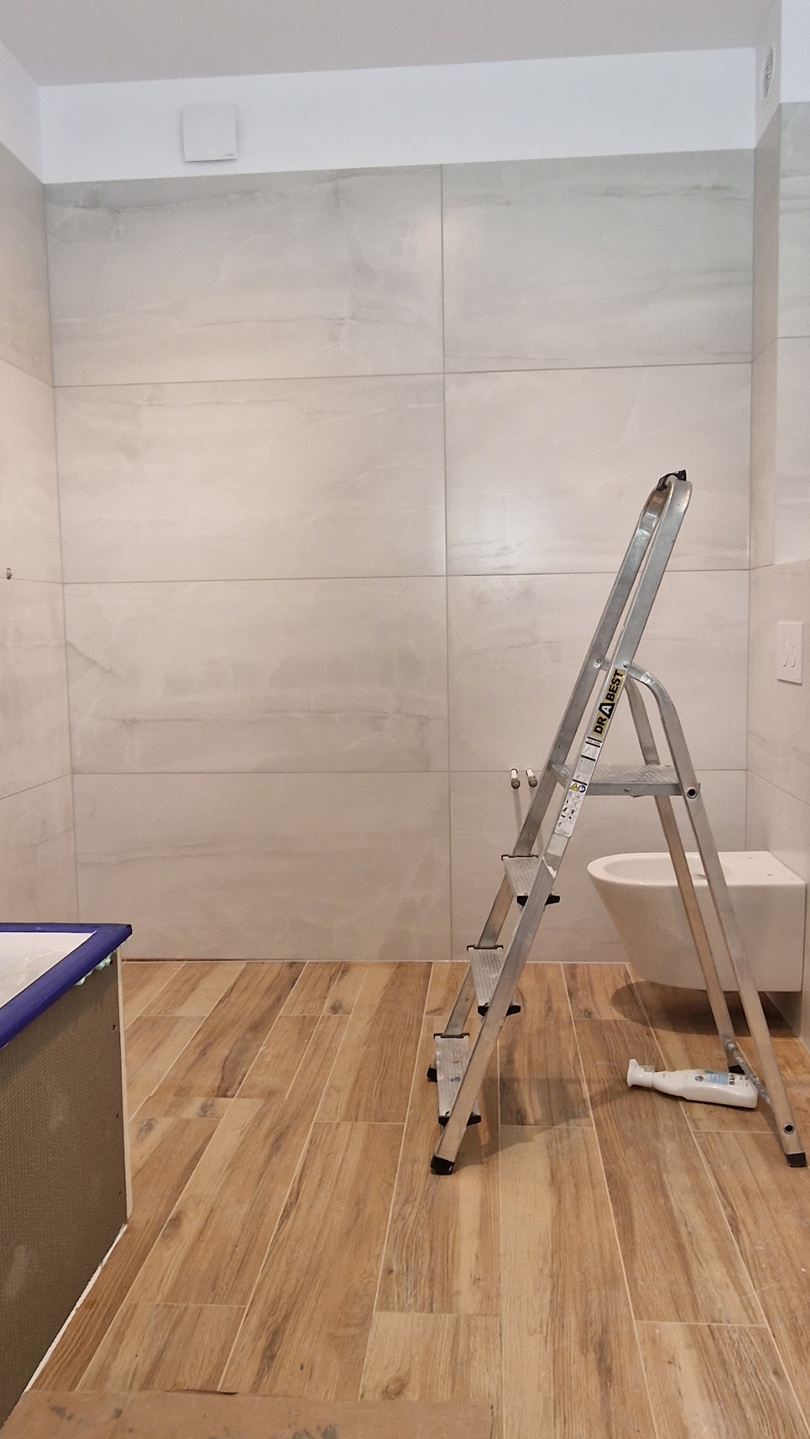 Łazienka w trakcie wykańczania: jasne płytki na ścianach, drewnopodobna podłoga, drabina aluminiowa, częściowo zabudowana wanna, widoczna toaleta.