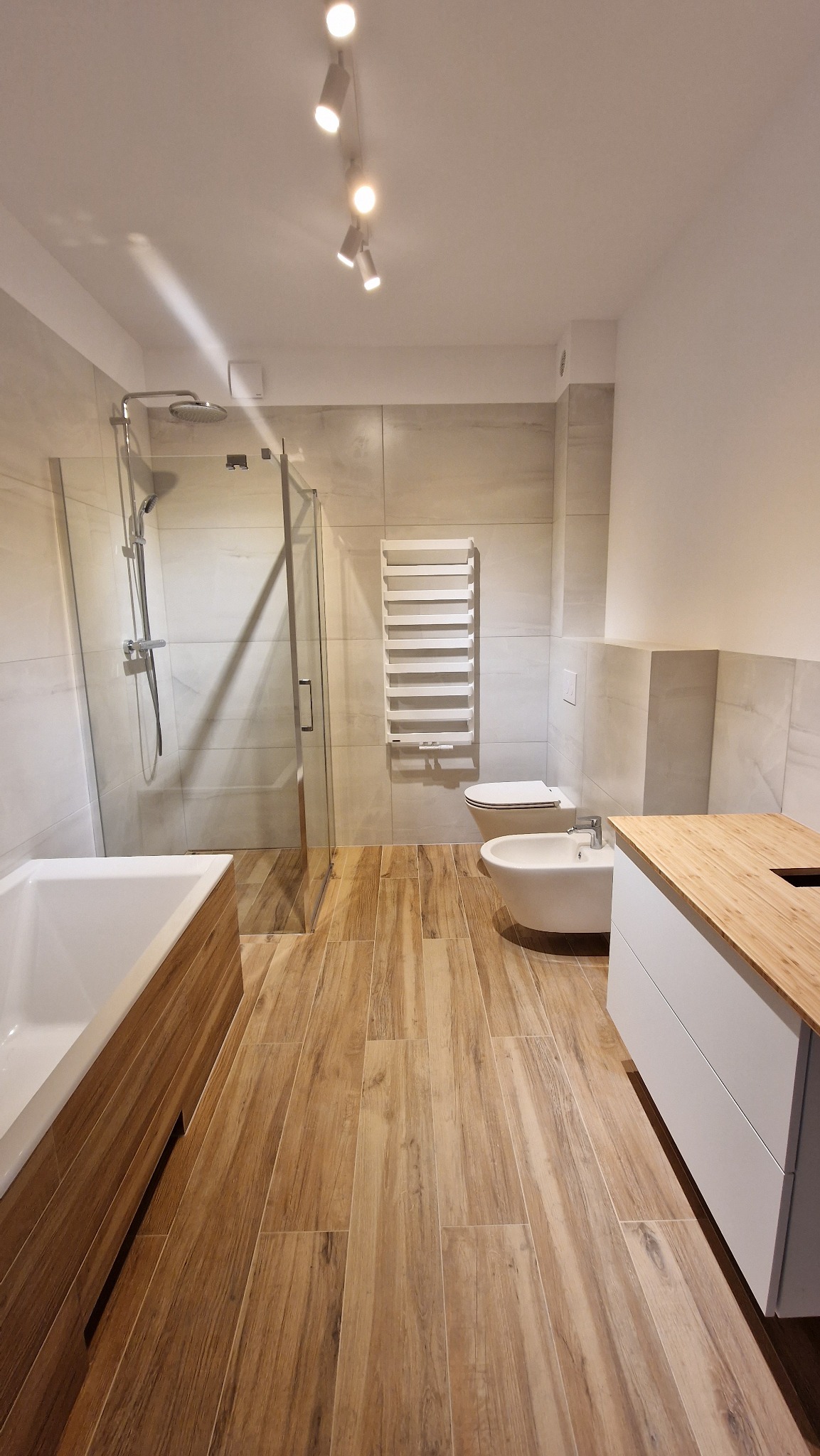 Nowoczesna łazienka z płytkami imitującymi drewno na podłodze, kabiną prysznicową ze szklaną ścianką, grzejnikiem drabinkowym, białą armaturą i blatem z jasnego drewna.