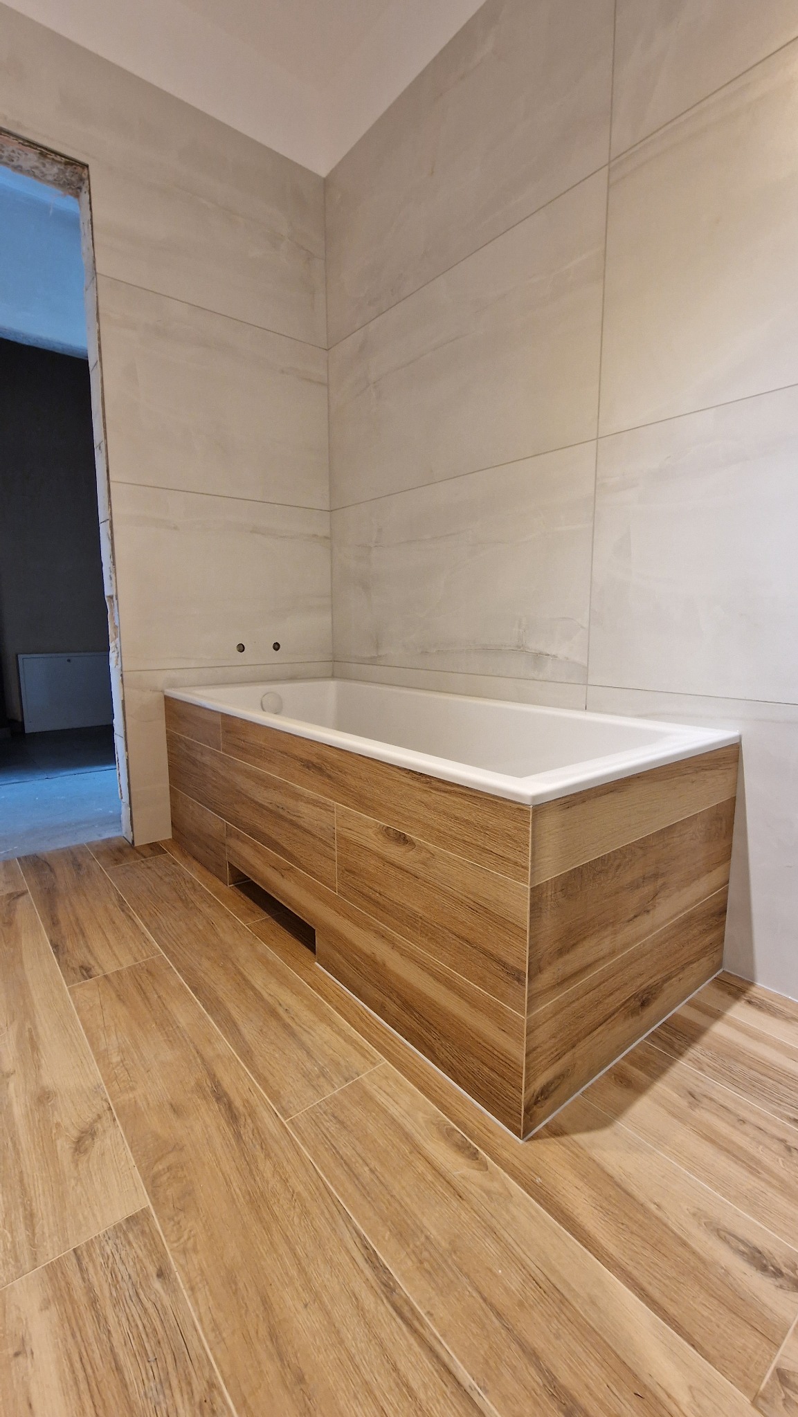 Wykończona łazienka z wanną obudowaną drewnopodobnymi panelami, ściany wyłożone jasnymi, dużymi płytkami, podłoga z drewnopodobnych płytek ułożonych wzdłużnie, widoczne dwa otwory na baterię wannową.