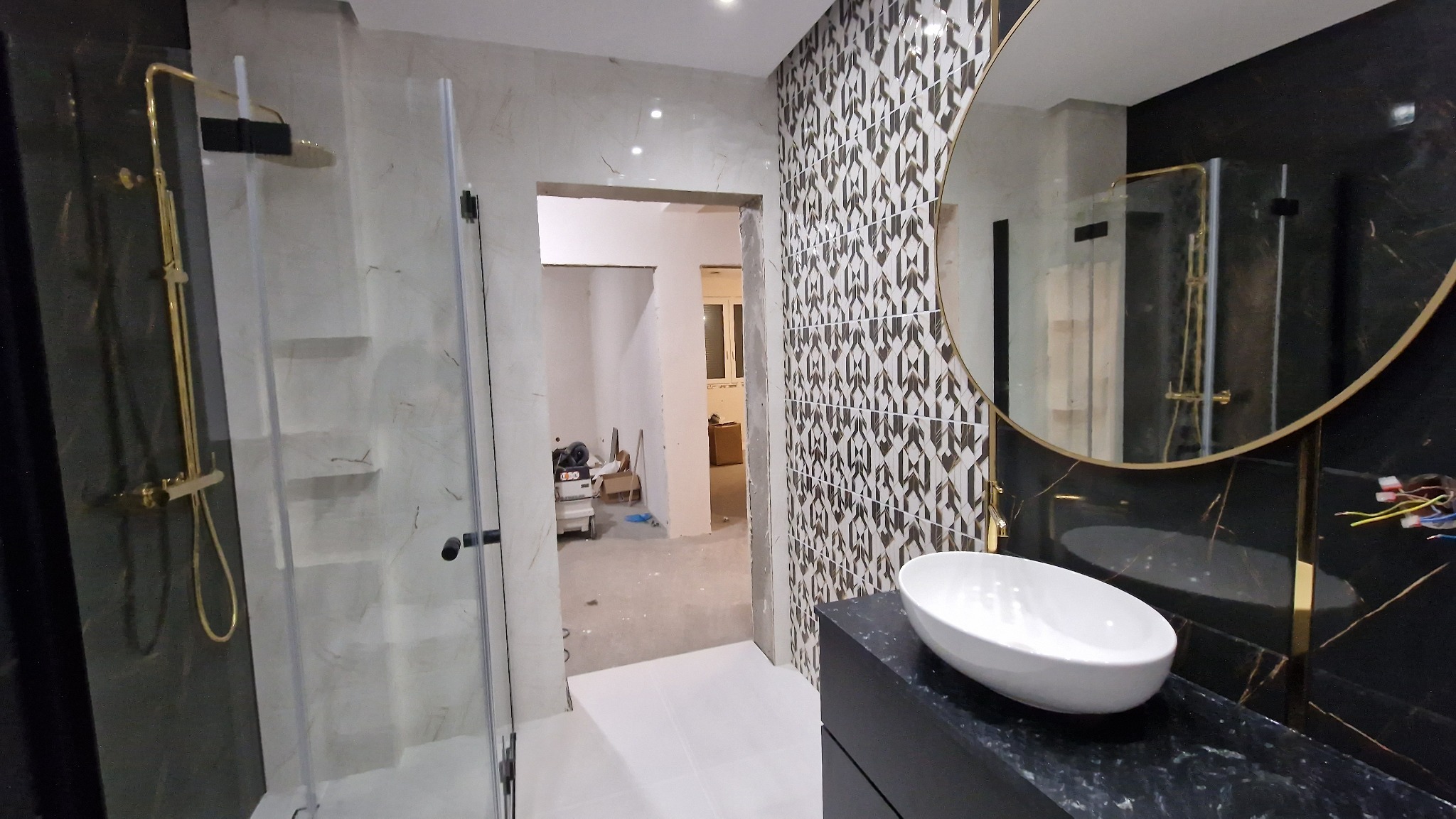 Wnętrze łazienki w trakcie wykańczania z widoczną kabiną prysznicową z złotą armaturą, umywalką nablatową i dekoracyjnymi płytkami na ścianie.
