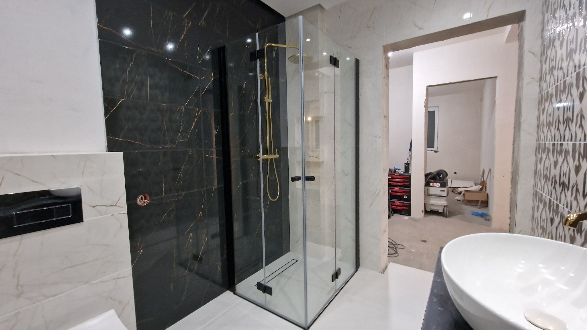 Nowoczesna kabina prysznicowa z czarnymi detalami i złotą armaturą na tle ściany z płytek imitujących czarny marmur z żyłami, widok na niedokończone pomieszczenie przez otwarte drzwi.
