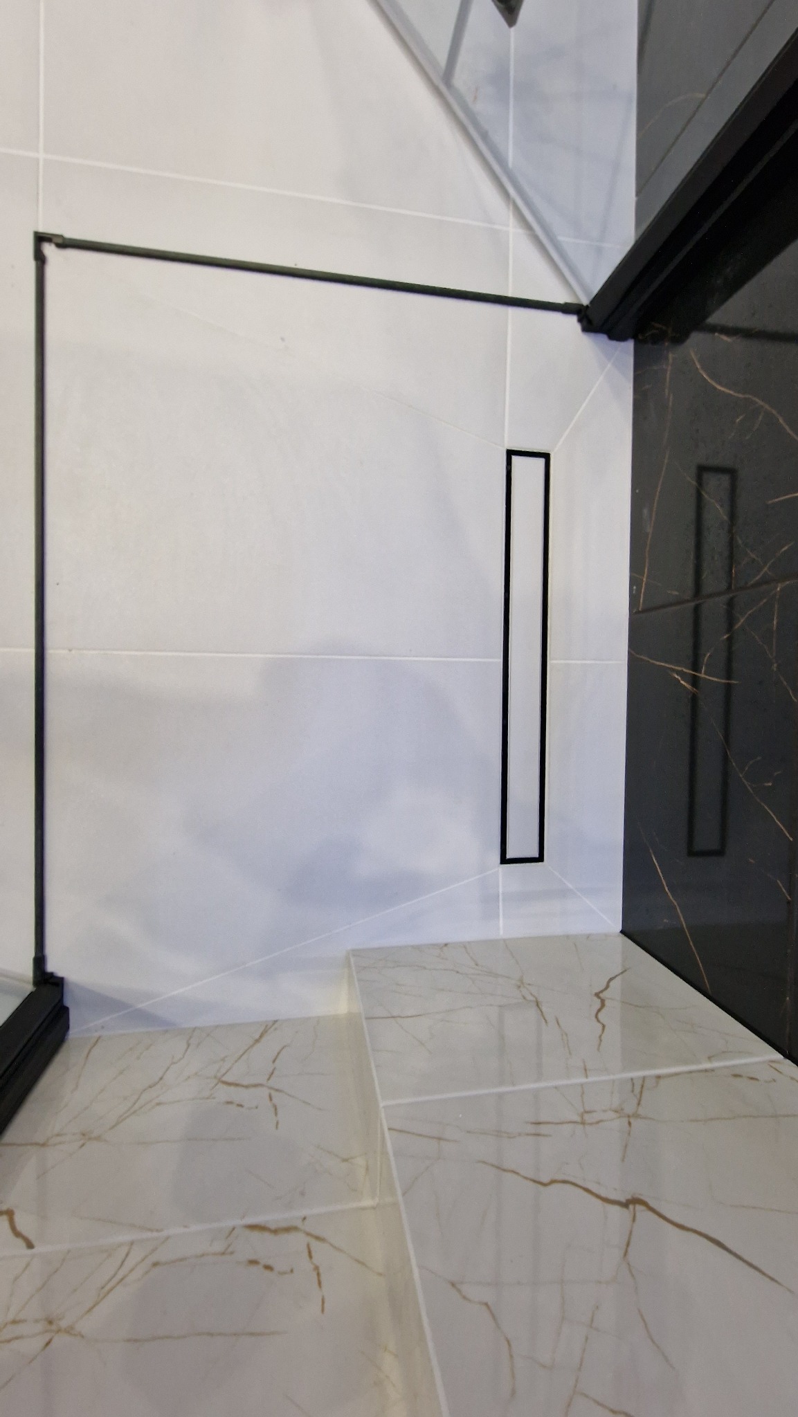 Kabina prysznicowa z odpływem liniowym w podłodze wyłożonej płytkami imitującymi marmur, ściany wyłożone jasnymi płytkami z czarną ramą kabiny.