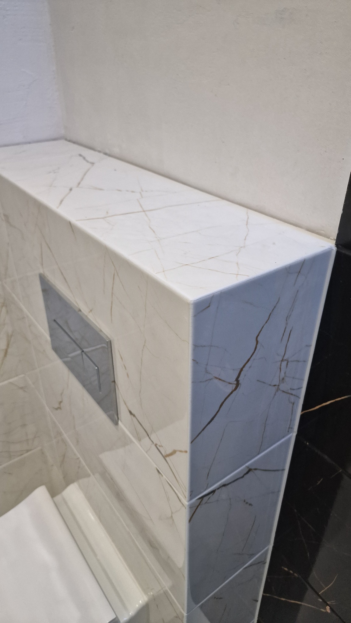 Obudowa stelaża podtynkowego WC wykończona płytkami imitującymi biały marmur z brązowymi żyłami, widoczny chromowany przycisk spłukujący i fragment miski ustępowej oraz czarne płytki na ścianie.