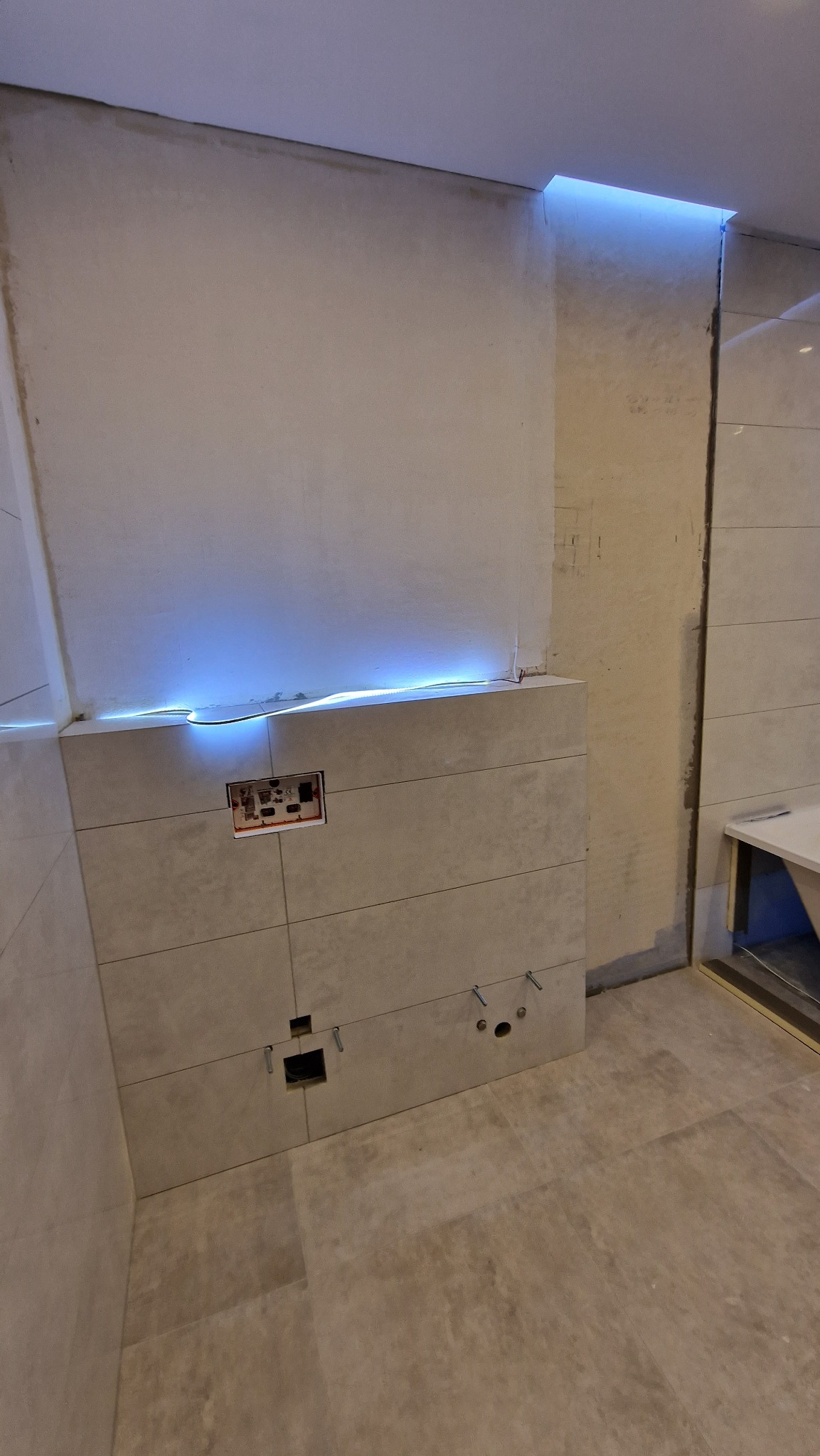 Montaż płytek ceramicznych w łazience z podświetleniem LED nad zabudową WC, widoczne otwory na instalację sanitarną i puszkę elektryczną.