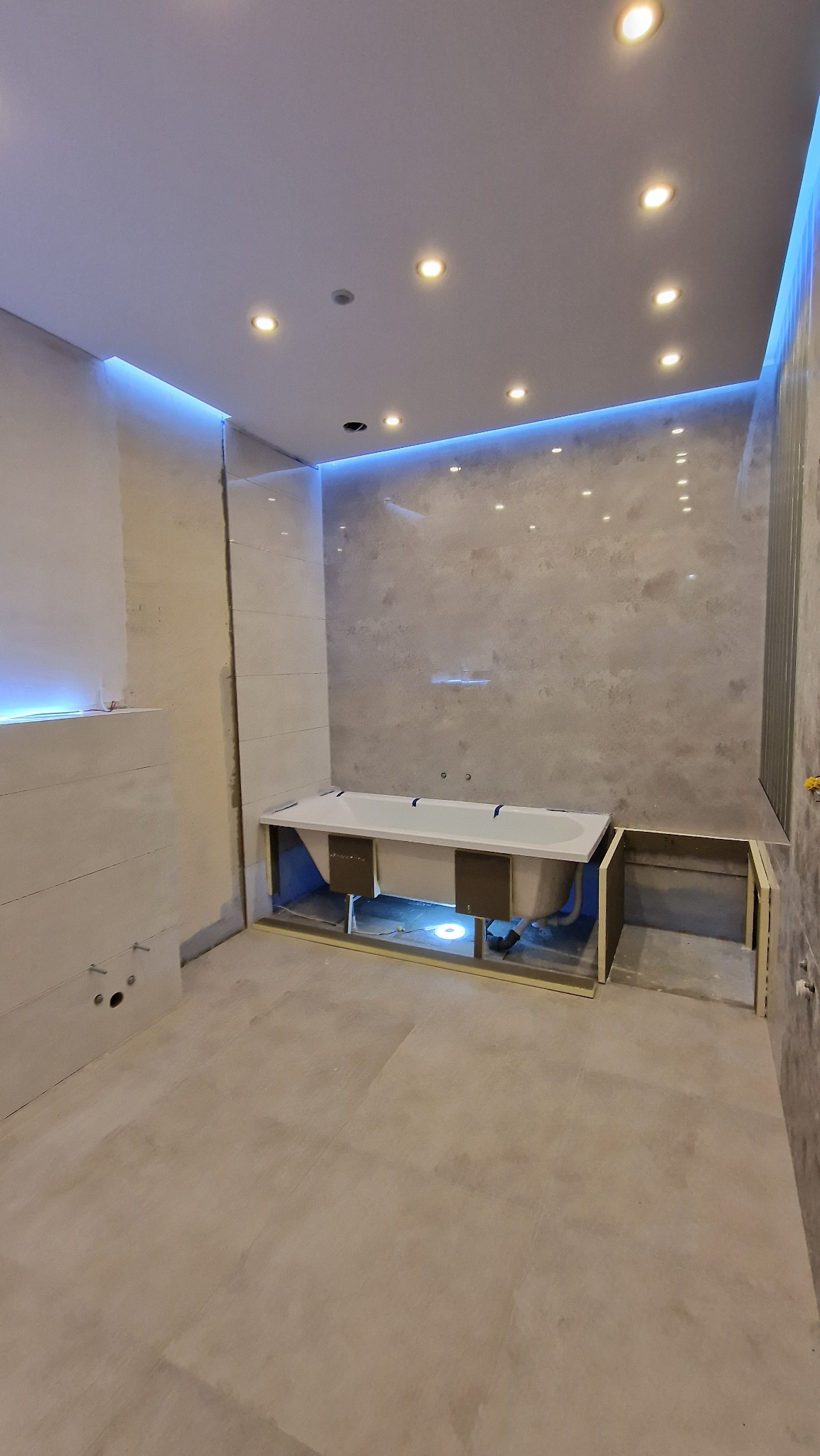 Łazienka w trakcie wykańczania: obudowana wanna, częściowo położone płytki ścienne, podświetlenie LED sufitu i ścian, widoczne instalacje hydrauliczne.