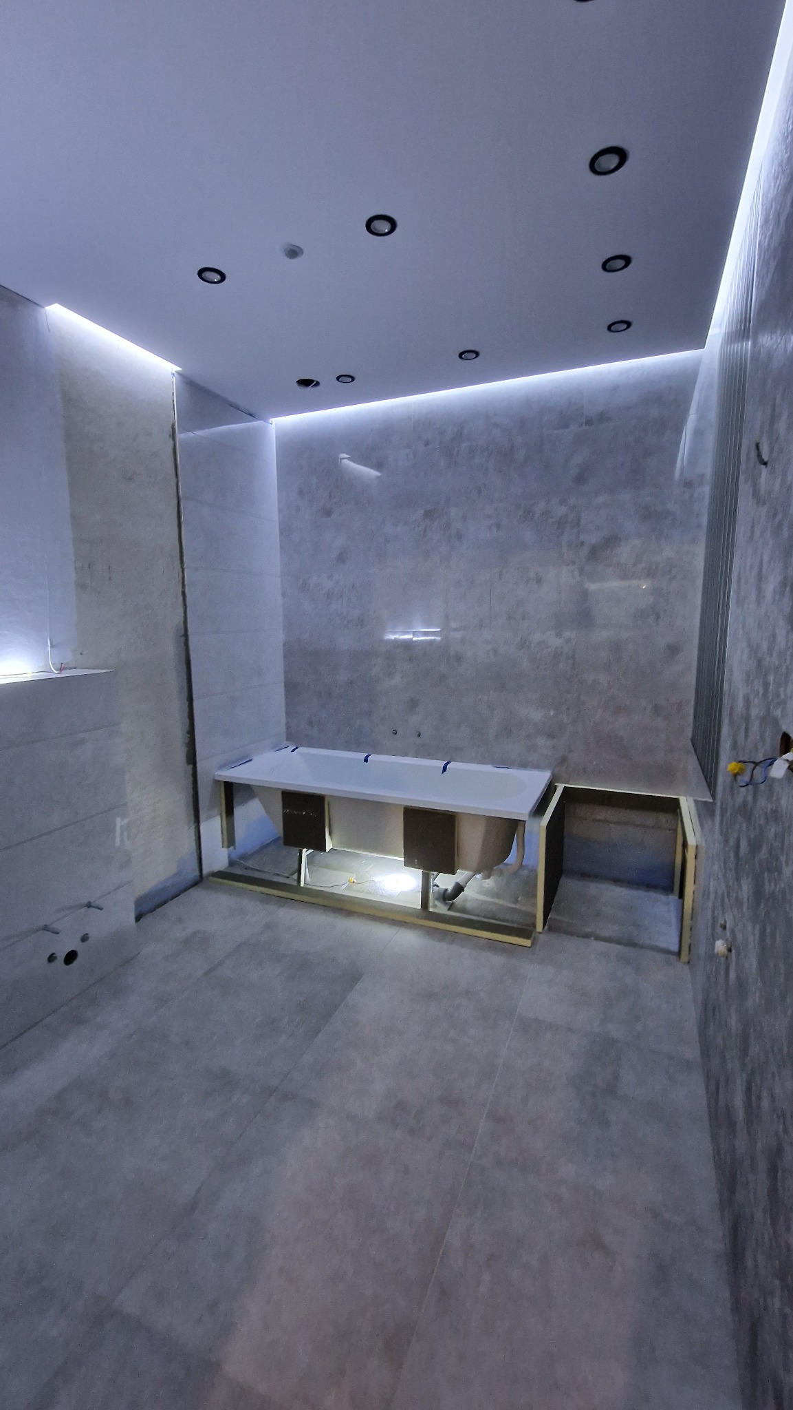 Łazienka w trakcie remontu, widoczna zabudowana wanna z szarą obudową, szare płytki na ścianach i podłodze, oraz podwieszany sufit z oświetleniem punktowym i listwą LED.