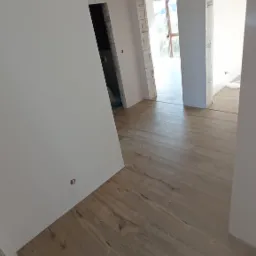 Wnętrze w trakcie wykańczania: świeżo ułożona podłoga z paneli imitujących drewno, białe ściany po szpachlowaniu, widoczne otwory drzwiowe w surowym stanie.