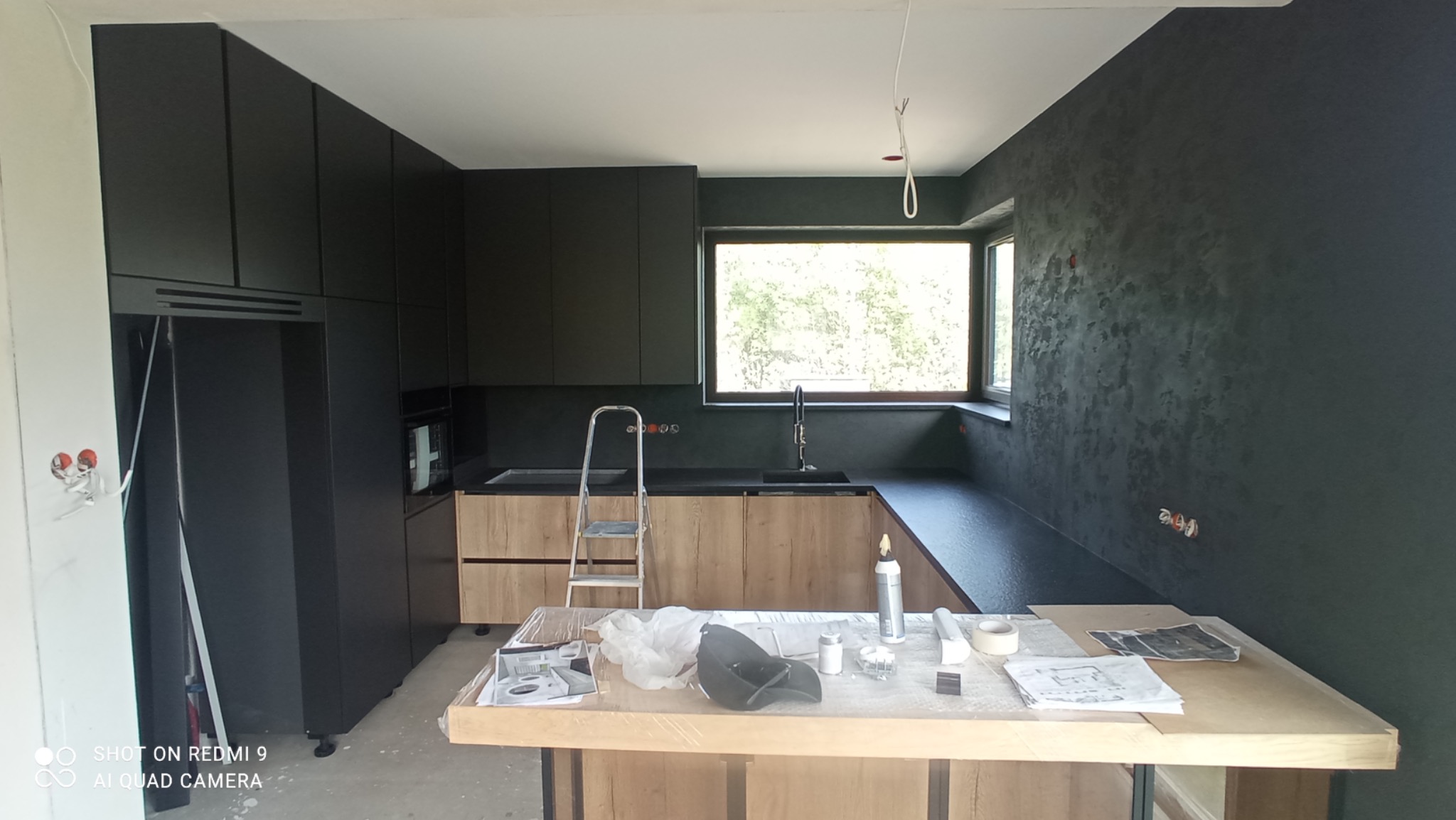 Częściowo wykończona, nowoczesna kuchnia w Radomiu z czarnymi szafkami i drewnianymi frontami, widoczna drabina i materiały wykończeniowe na blacie wyspy.