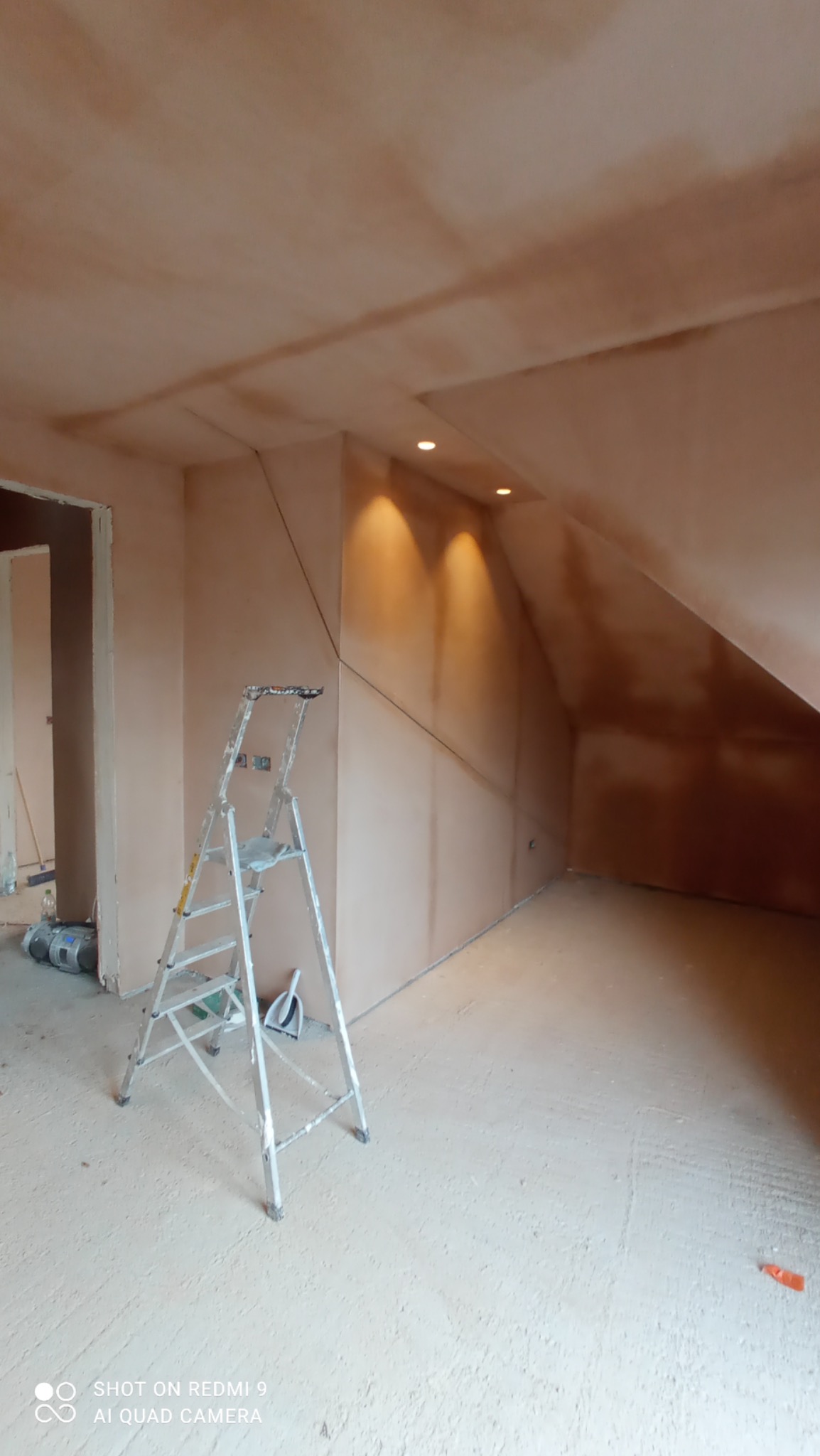 Pokój na poddaszu po nałożeniu pierwszej warstwy gładzi gipsowej na ściany i sufit, widoczna aluminiowa drabina oraz oświetlenie punktowe w suficie.