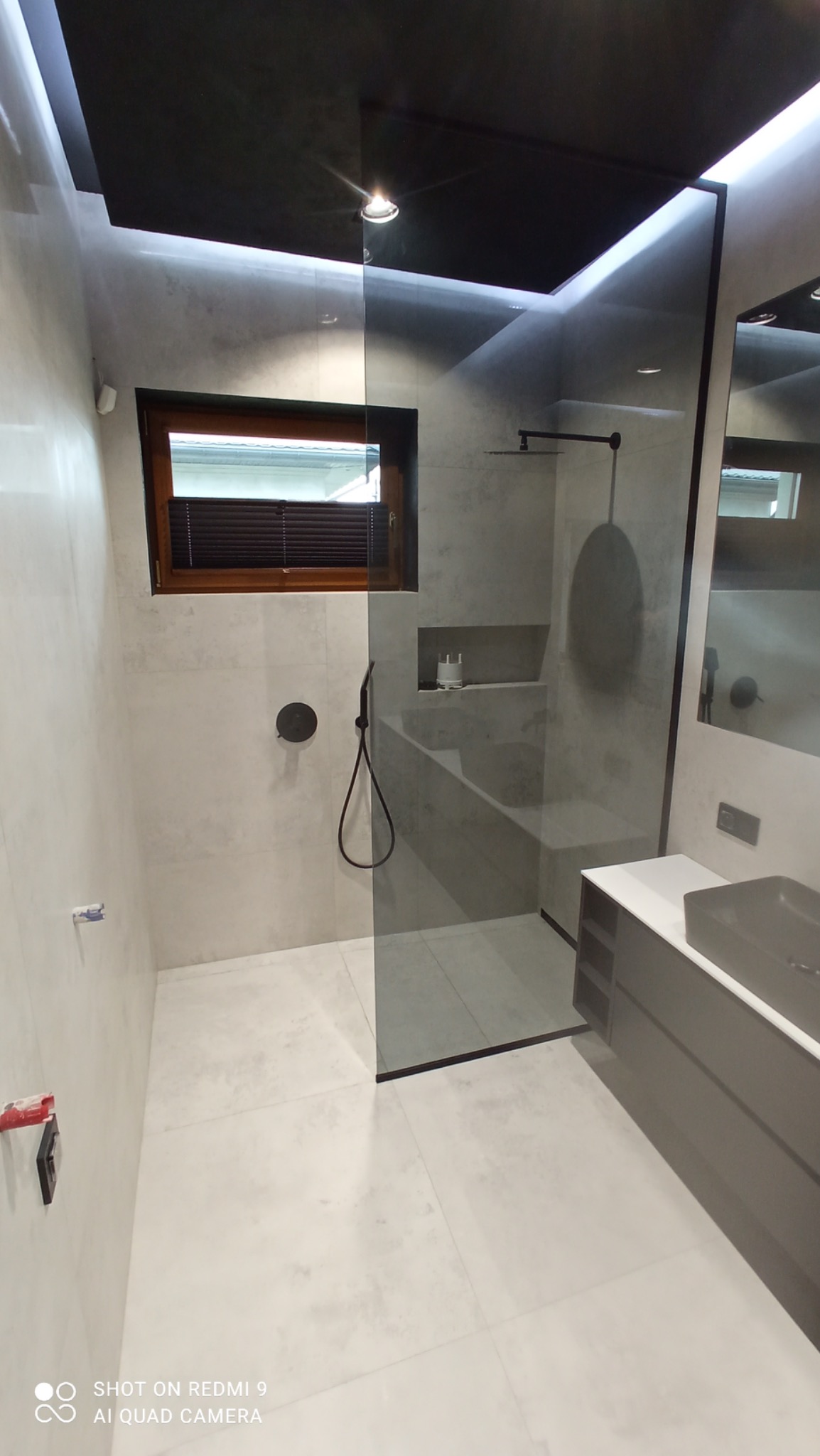 Nowoczesna łazienka z szarą armaturą, kabiną prysznicową typu walk-in, oknem i czarnym sufitem podwieszanym z oświetleniem punktowym.