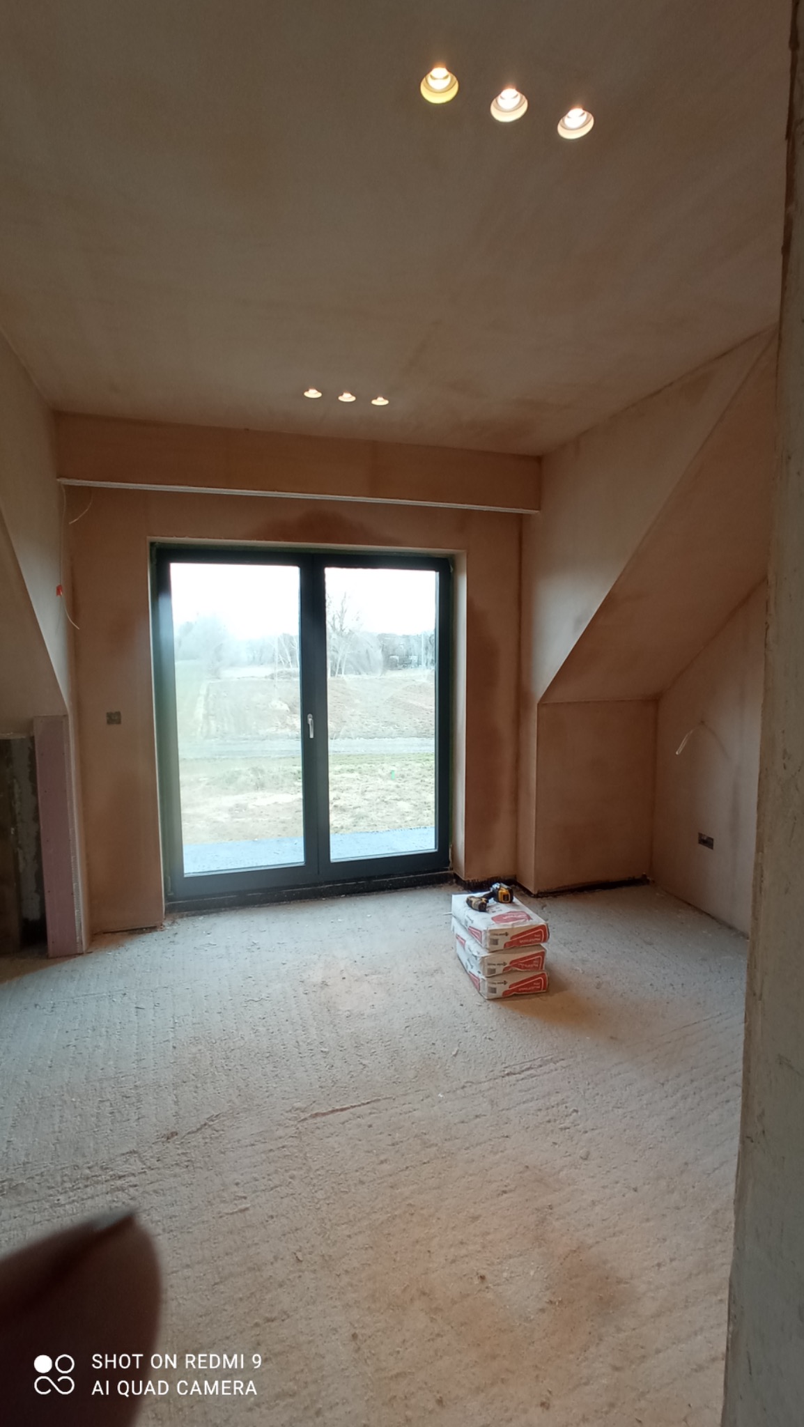 Pokój w trakcie wykańczania, świeżo otynkowane ściany w beżowym odcieniu, widoczne puszki z zaprawą na podłodze, okno balkonowe z widokiem na zewnątrz, sufit z oświetleniem punktowym.