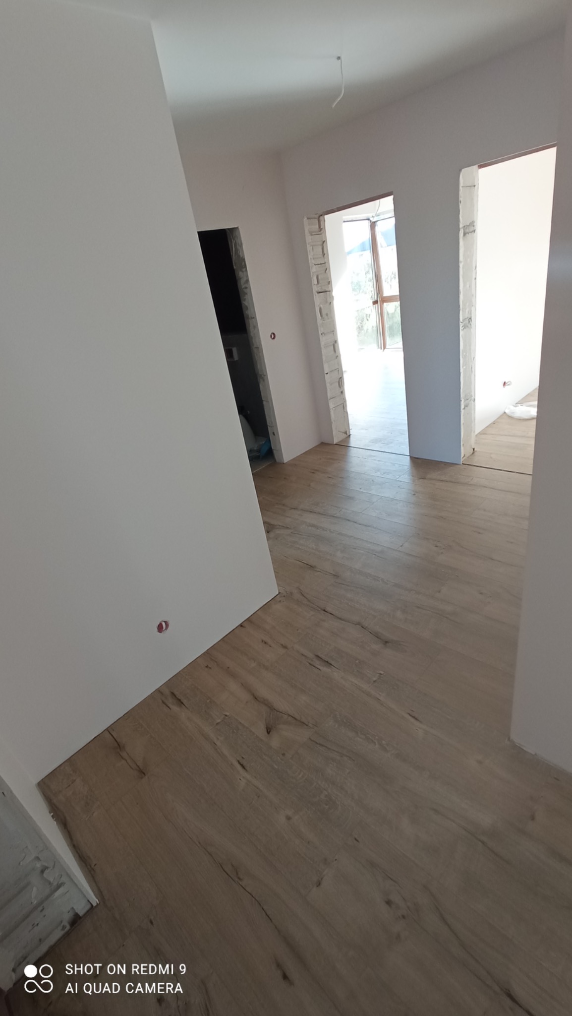 Wnętrze w trakcie wykańczania: świeżo ułożona podłoga z paneli imitujących drewno, białe ściany po szpachlowaniu, widoczne otwory drzwiowe w surowym stanie.