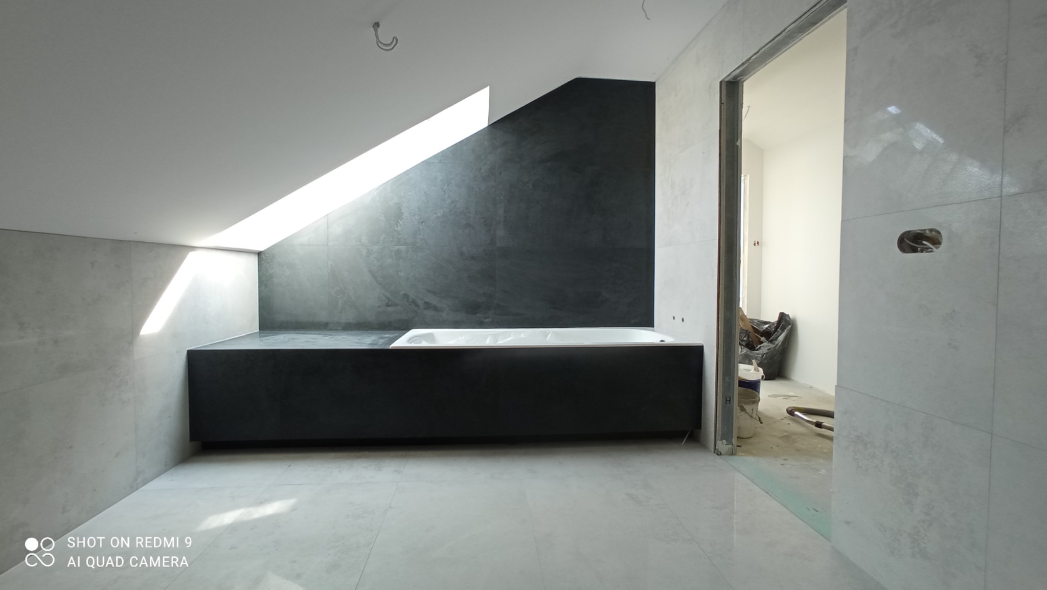 Wnętrze łazienki w trakcie wykańczania z wanną w zabudowie, szare płytki na ścianach i podłodze, okno dachowe w skosie, widoczne ślady prac wykończeniowych i niedokończone otwory na gniazdka.