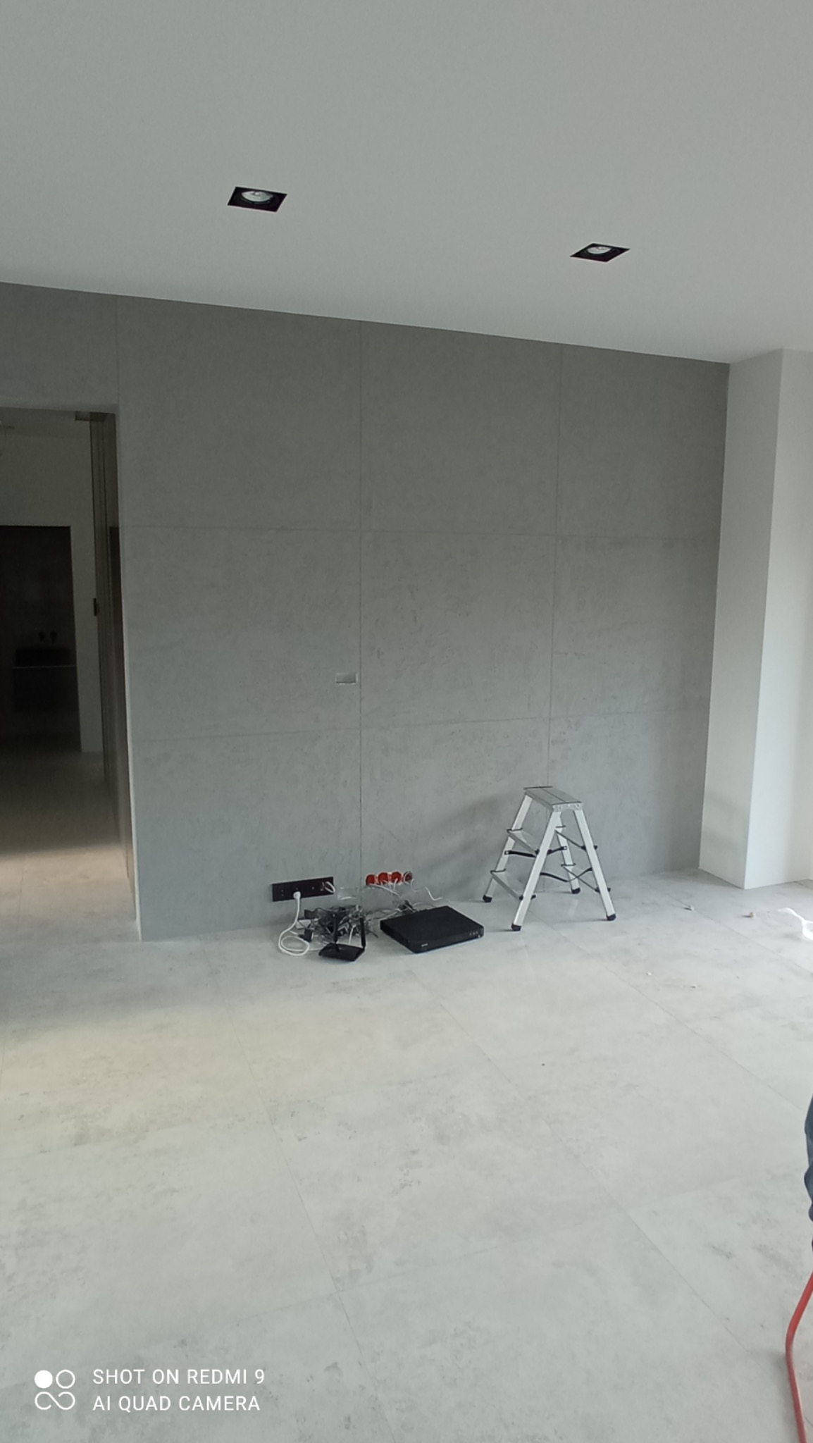 Ściana wykończona płytami imitującymi beton, z widocznymi gniazdkami elektrycznymi i przewodami, drabina aluminiowa w pokoju podczas prac wykończeniowych, oświetlenie sufitowe punktowe.