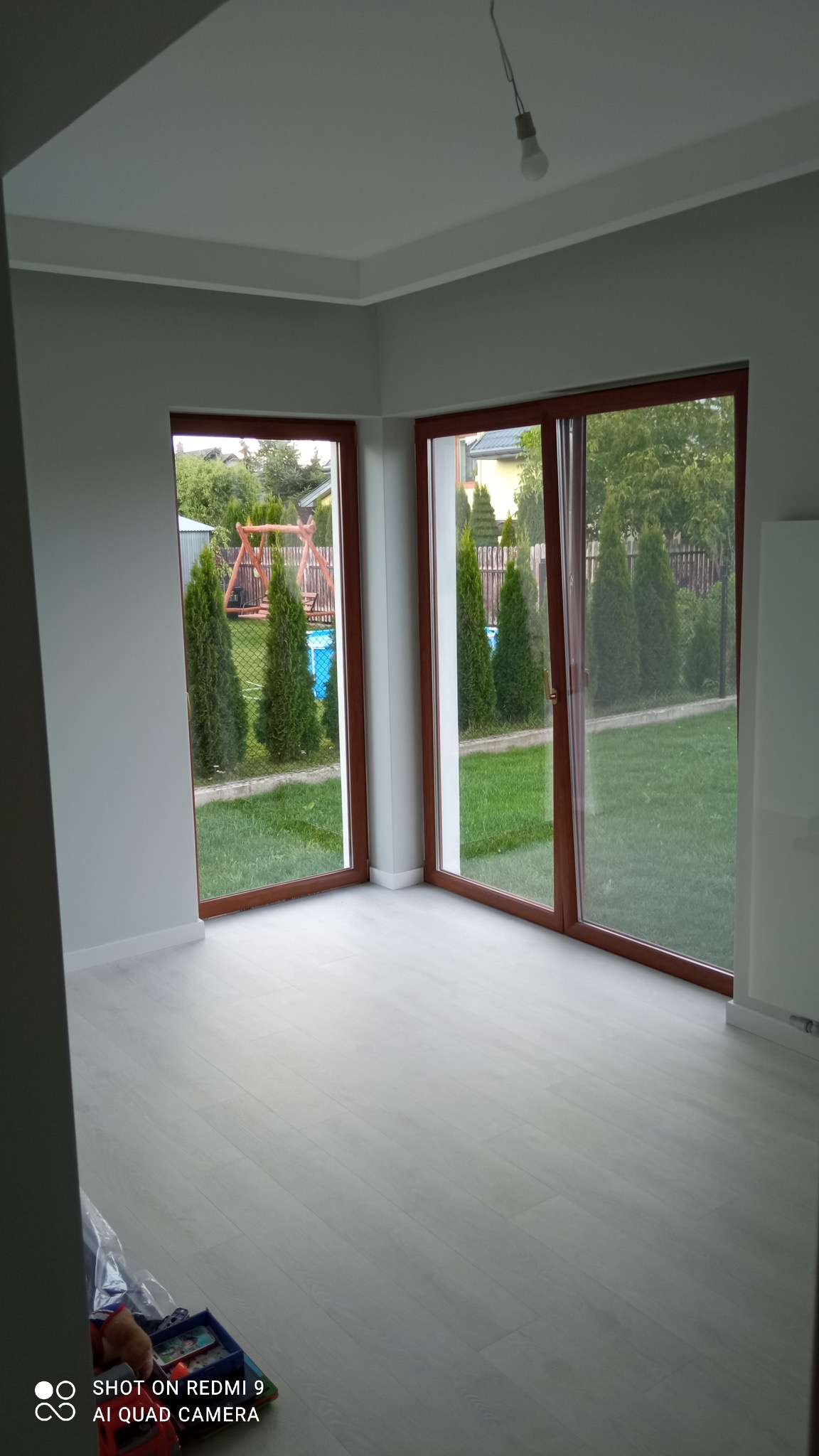 Jasne, odnowione wnętrze z drewnianymi oknami przesuwnymi, świeżo pomalowane ściany w odcieniach szarości i jasna podłoga. Widok na zielony ogród z tujami i huśtawką przez okna.