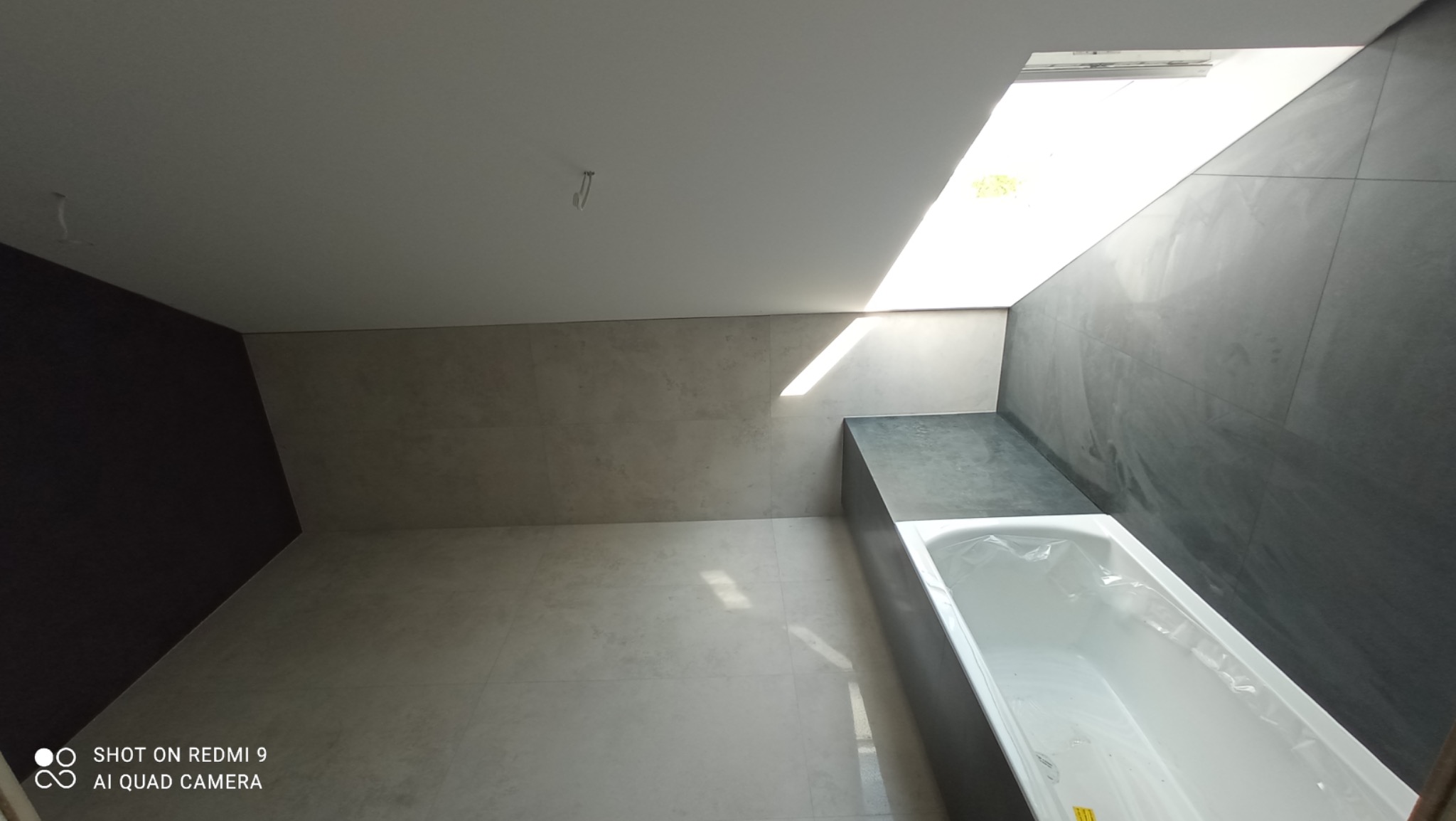 Nowoczesna łazienka z szarymi płytkami, białą wanną zabezpieczoną folią i oknem dachowym wpuszczającym jasne światło.