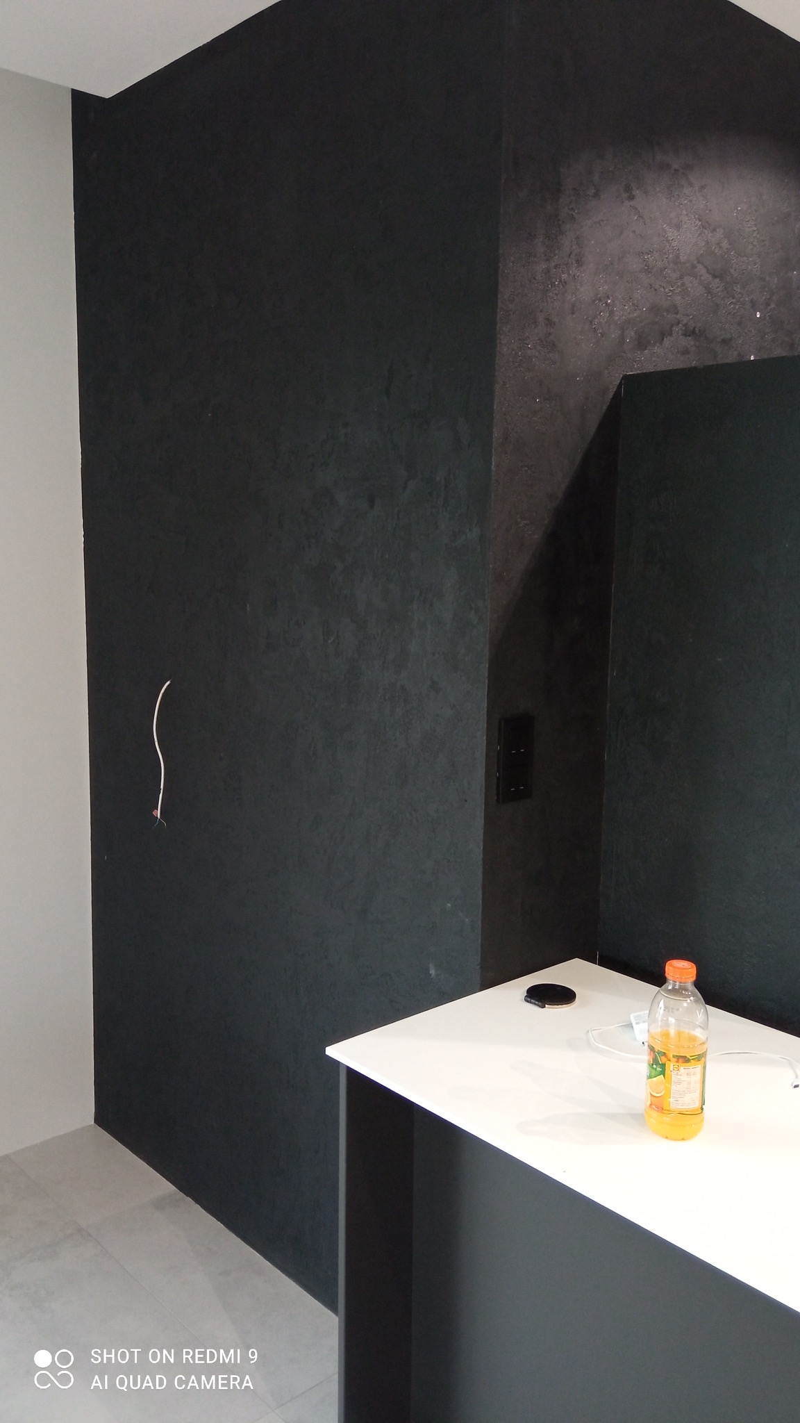 Ciemna, teksturowana ściana z widoczną instalacją elektryczną i gniazdkiem, biały blat z butelką soku pomarańczowego, szara podłoga z płytek.