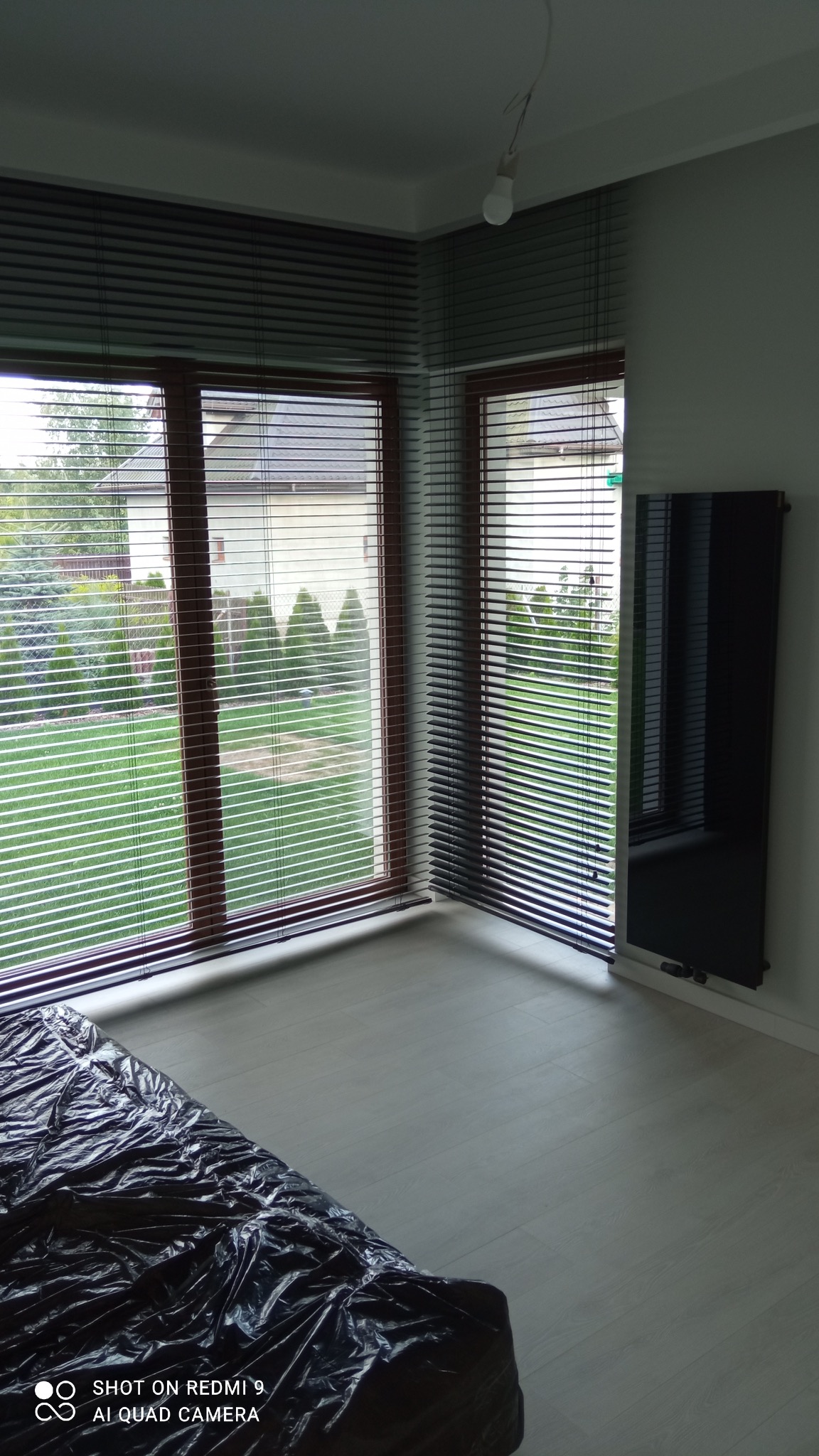 Pokój po remoncie z nową, jasną podłogą, osłoniętą folią, oraz zamontowanymi drewnianymi oknami z żaluzjami i czarnym grzejnikiem na ścianie.