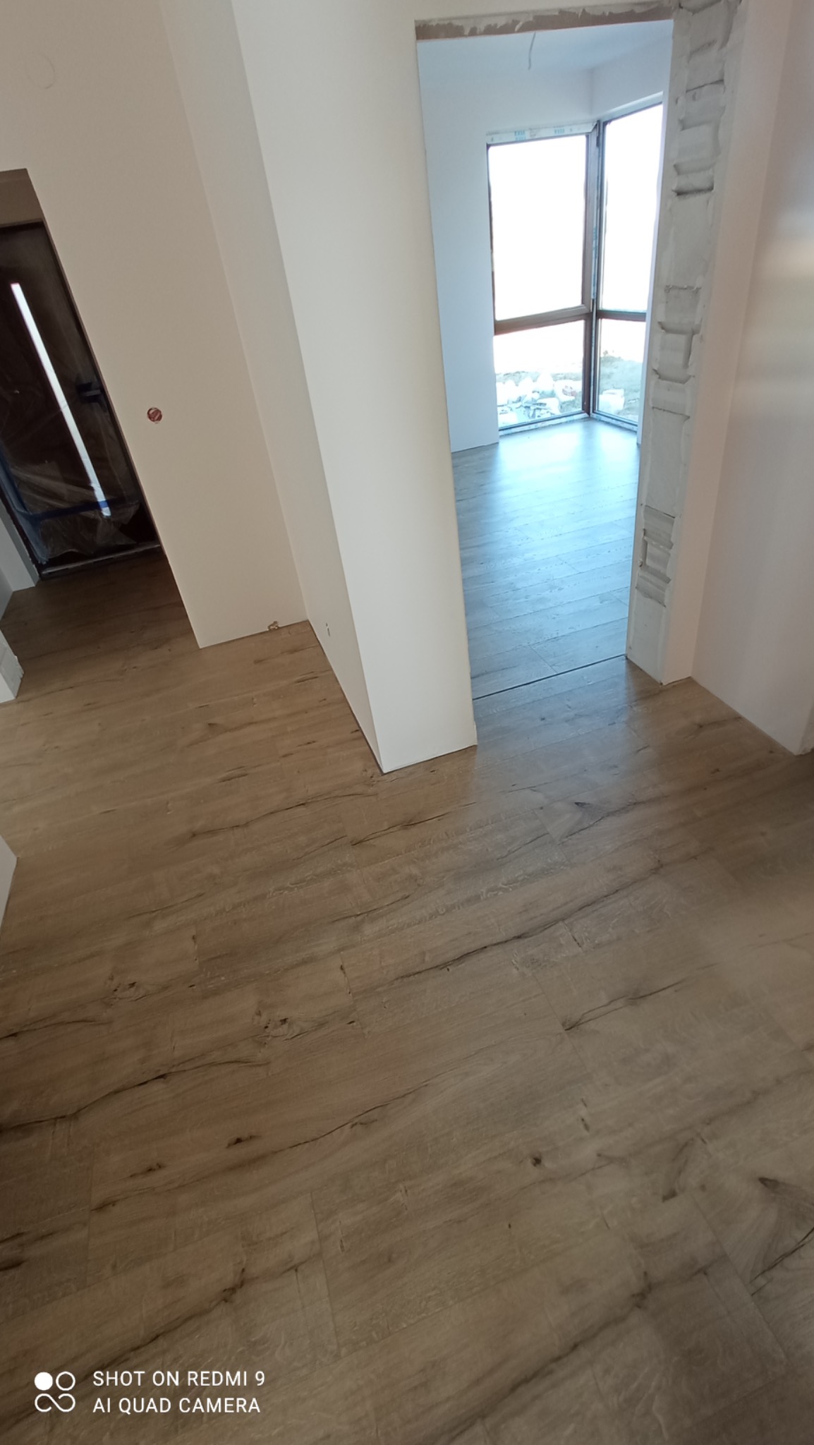 Ułożona podłoga z paneli o wzorze drewna w jasnym odcieniu, widok na drzwi i okno w trakcie prac wykończeniowych, surowe ściany w tle.