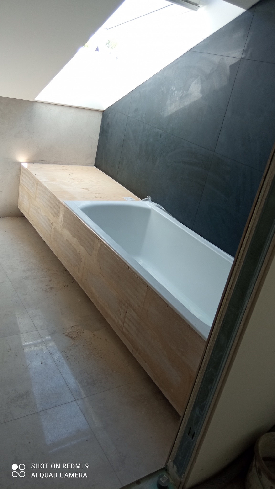 Biała wanna obudowana surowym drewnem, umieszczona pod oknem dachowym w łazience z ciemnymi płytkami na ścianie, widoczne ślady prac wykończeniowych na podłodze.