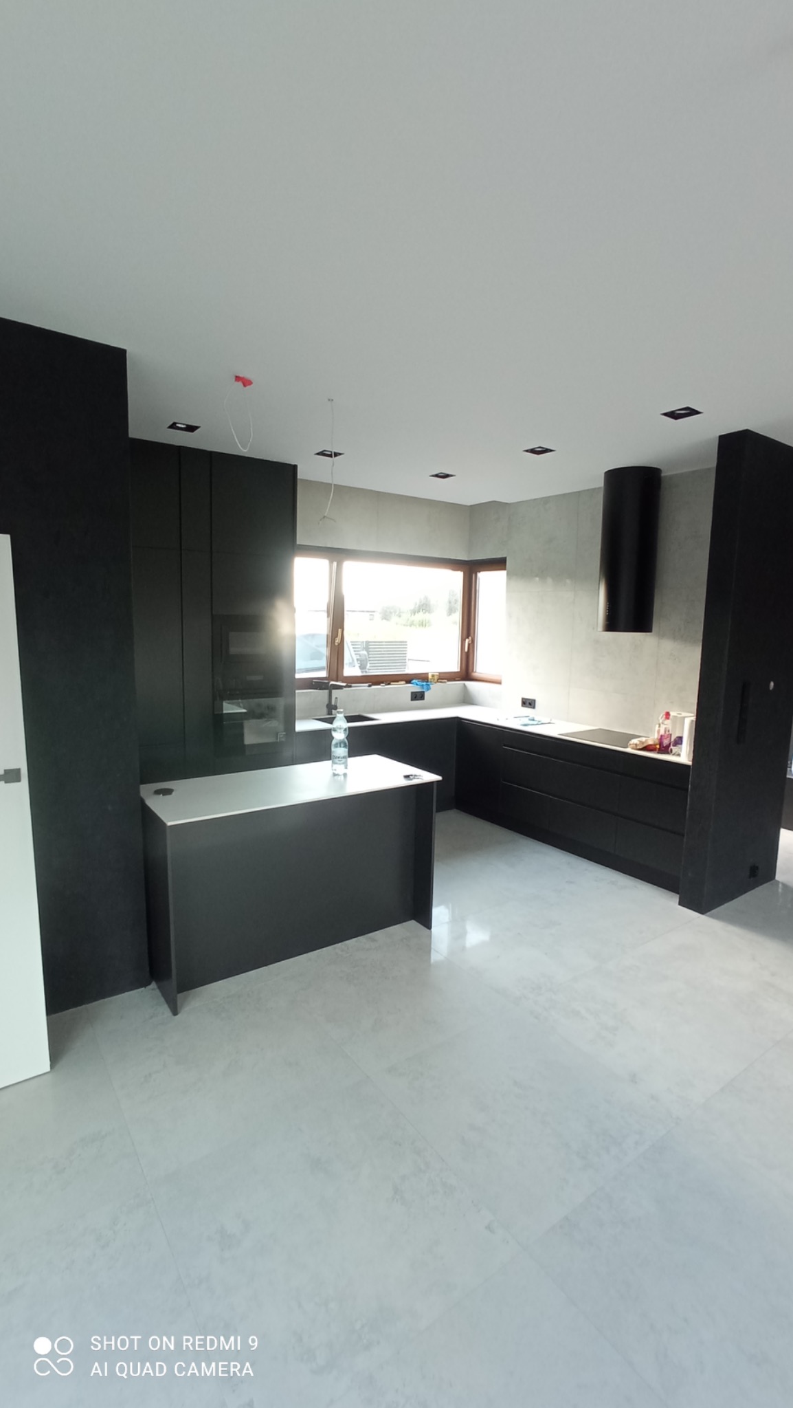 Nowoczesna, minimalistyczna kuchnia w odcieniach szarości i czerni z wyspą, jasnymi płytkami podłogowymi i oknem w tle; widoczne niedokończone podłączenie oświetlenia sufitowego.