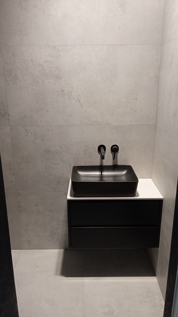 Nowoczesna łazienka z czarną, kwadratową umywalką nablatową i minimalistyczną szafką pod umywalką, szare płytki wielkoformatowe na ścianach i podłodze, czarna armatura.
