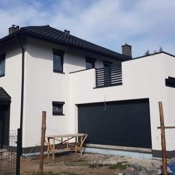 Move House Sp. Z o.o. - Nowoczesny, dwukondygnacyjny dom z garażem w bryle, wykończony na biało z czarnymi akcentami, balkon z czarną balustradą, widok z zewnątrz, w tle błękitne niebo.