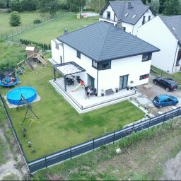 Move House Sp. Z o.o. - Nowoczesny, biały dom z ciemnym dachem, widok z lotu ptaka. Na trawniku basen, trampolina i plac zabaw. Na tarasie siedzą osoby. Zaparkowane samochody.