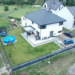 Nowoczesny, biały dom z ciemnym dachem, widok z lotu ptaka. Na trawniku basen, trampolina i plac zabaw. Na tarasie siedzą osoby. Zaparkowane samochody.