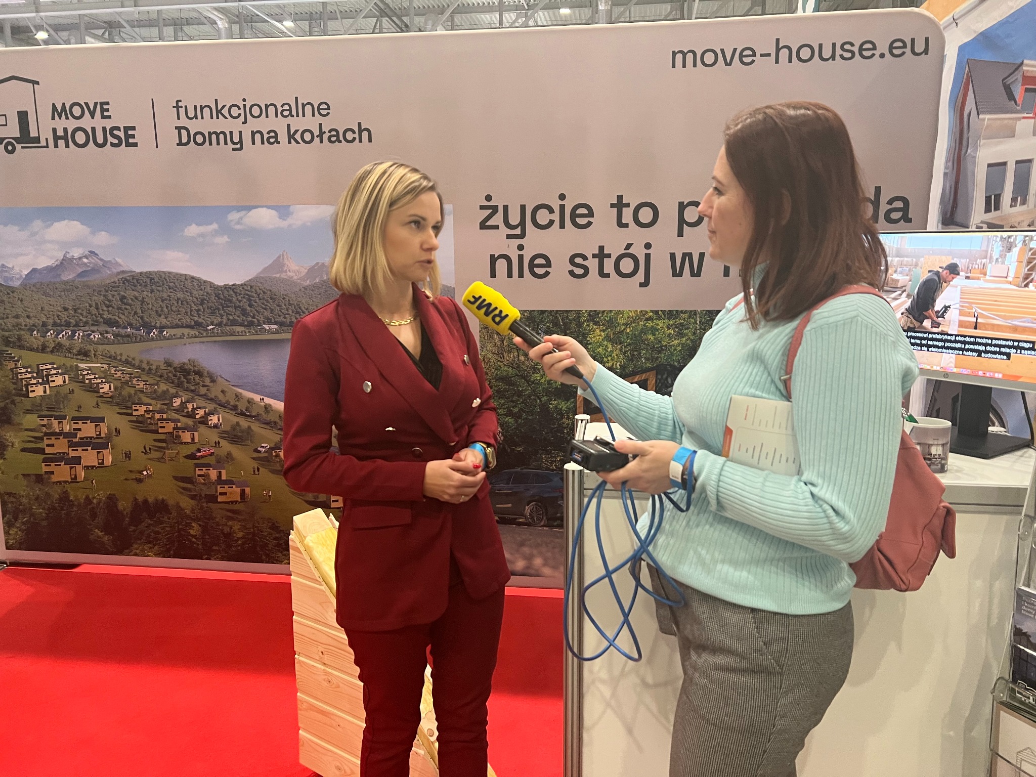 Wywiad na stoisku Move House: kobieta w czerwonym garniturze odpowiada na pytania dziennikarza RMF na tle grafiki z domami na kołach i budową domu szkieletowego.