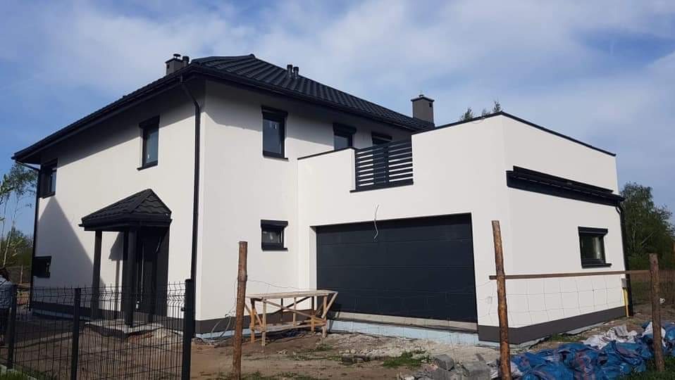 Nowoczesny, dwukondygnacyjny dom z garażem w bryle, wykończony na biało z czarnymi akcentami, balkon z czarną balustradą, widok z zewnątrz, w tle błękitne niebo.