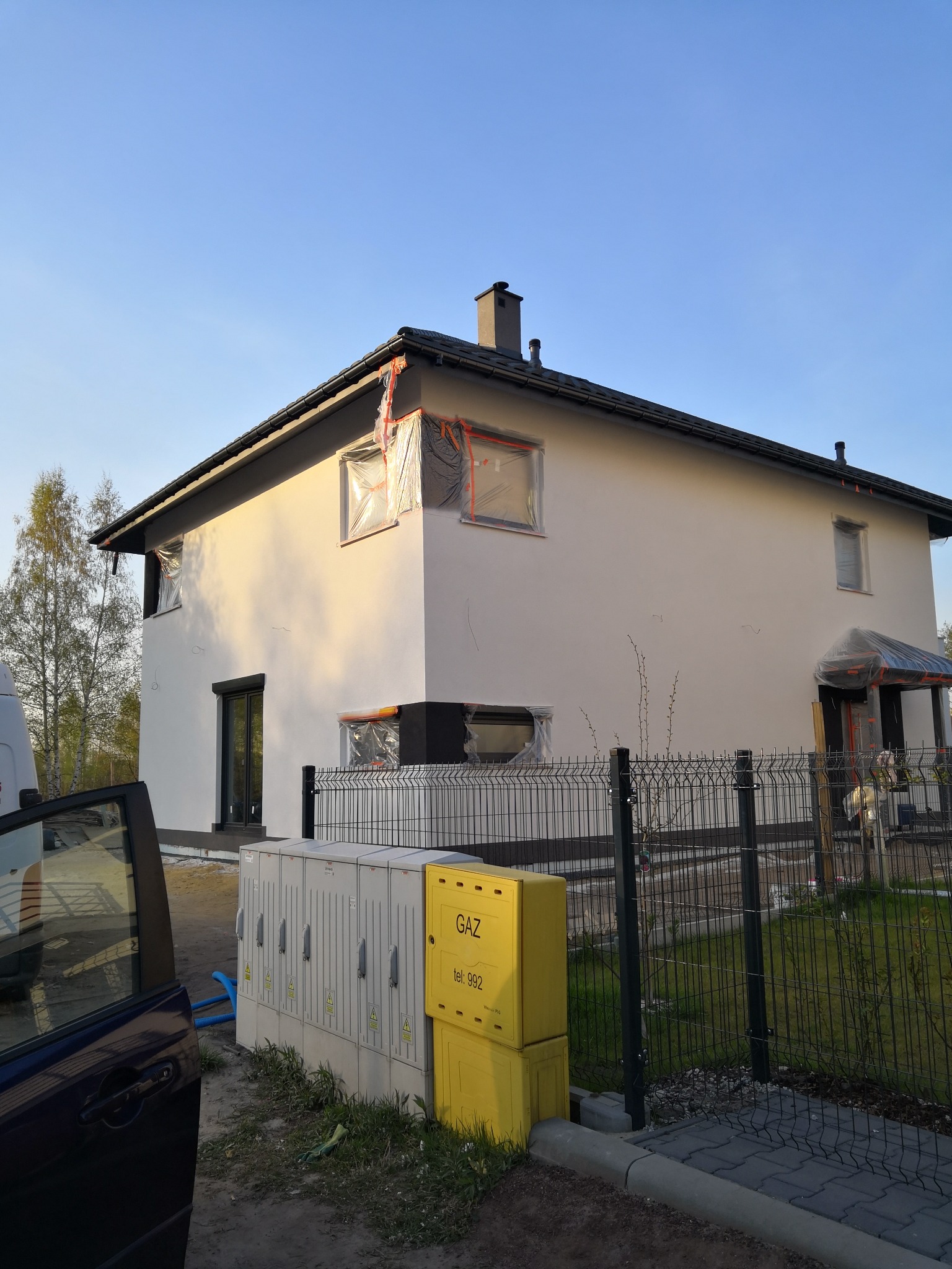 Dwukondygnacyjny dom z oknami zabezpieczonymi folią malarską, częściowo otynkowany, z czarną stolarką i dachem. Widoczne skrzynki gazowe i auto.