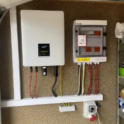 Biały falownik FOX ESS zamontowany na ścianie obok skrzynki rozdzielczej PV z widocznymi przewodami o różnych kolorach (czerwony, żółty, czarny, biały) oraz naklejką ostrzegawczą o napięciu...