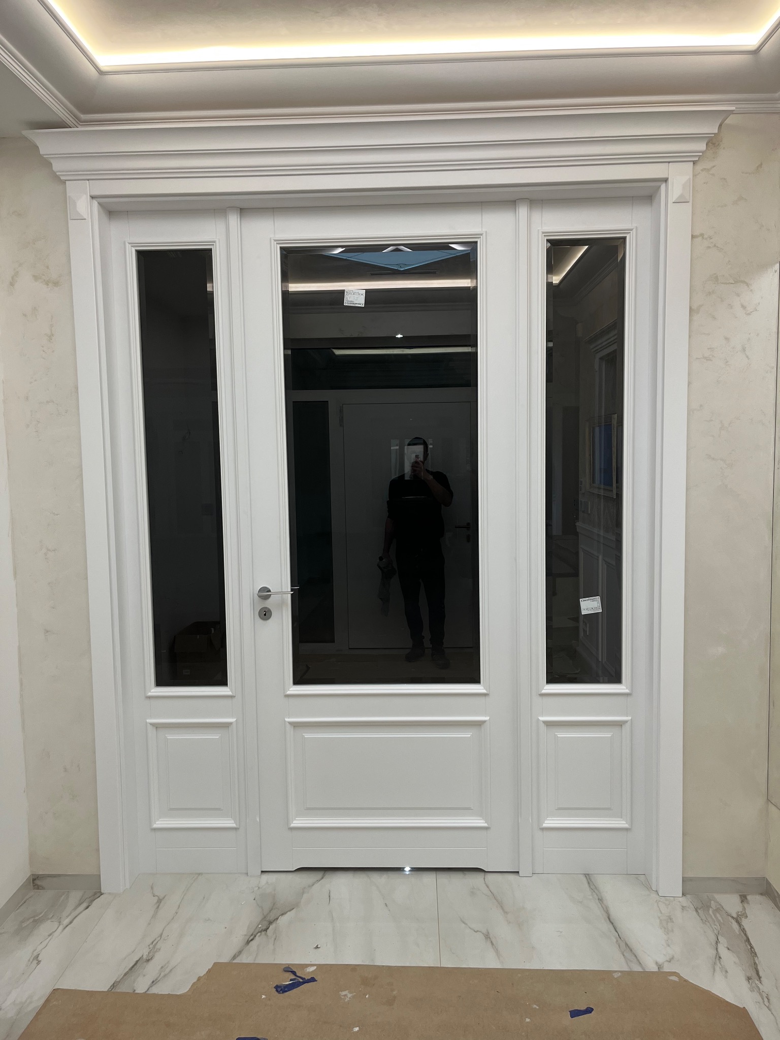 Nowoczesne, białe drzwi wewnętrzne z trzema szybami, widoczne odbicie osoby robiącej zdjęcie, marmurowe płytki podłogowe i jasne ściany z dekoracyjnym wykończeniem.